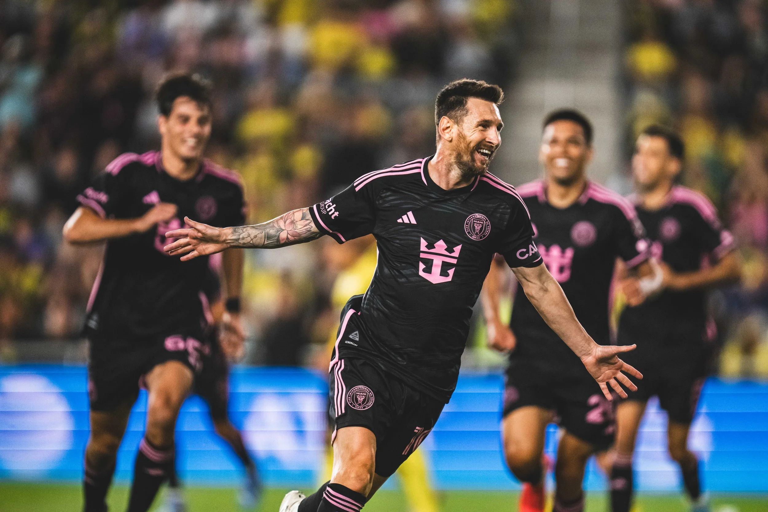 Soccer player celebrates a goal while teammates run behind him on the field, wearing black and pink uniforms.