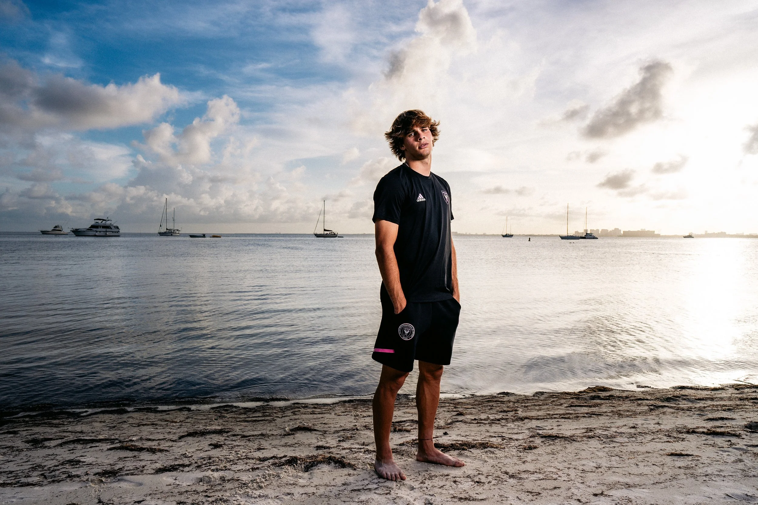 Young man standing barefoot on beach with hands in pockets, overlooking water with sailboats and yachts in the distance, under cloudy sky at sunset.