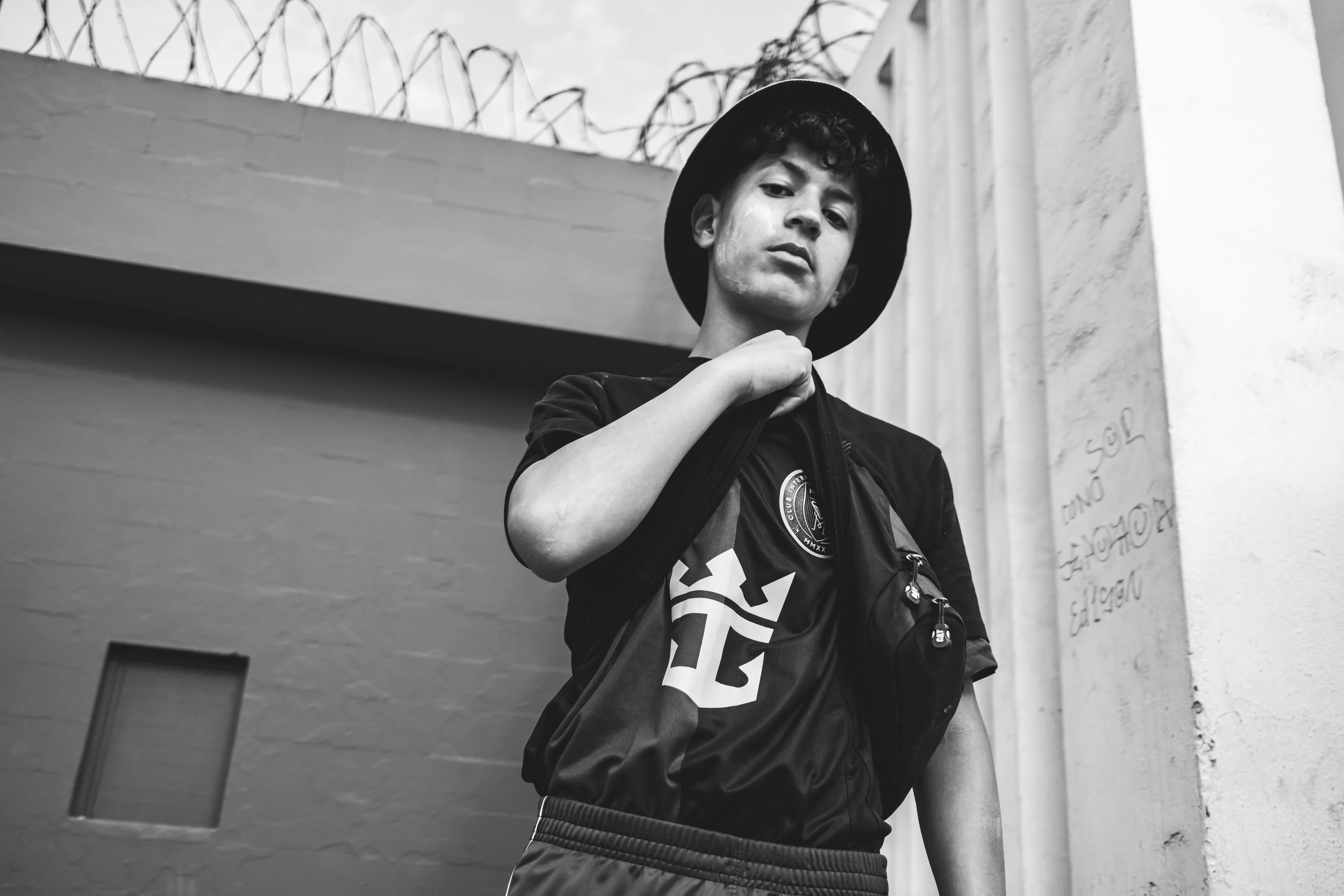 A young man with curly hair wearing a black hat and T-shirt, carrying a backpack, standing outdoors in front of a wall with graffiti and barbed wire.