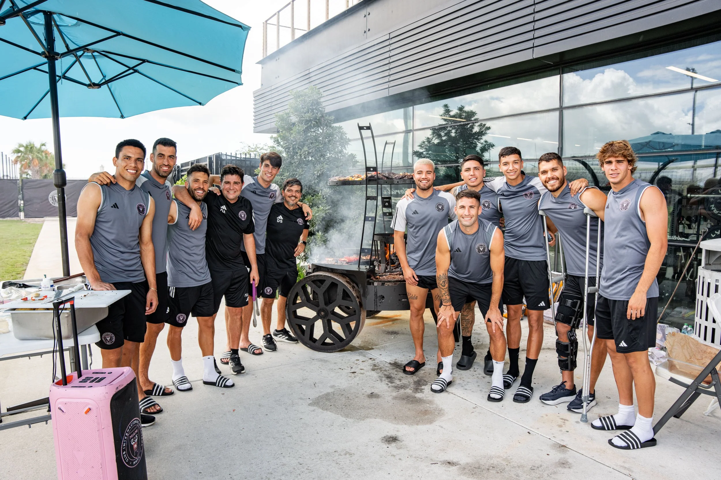 A group of mostly shirtless men in athletic shorts and slides, standing together outside near a building, smiling at the camera. One man has a crutch, and another has a knee brace. There is a grill with smoke and food cooking, with a pink suitcase an