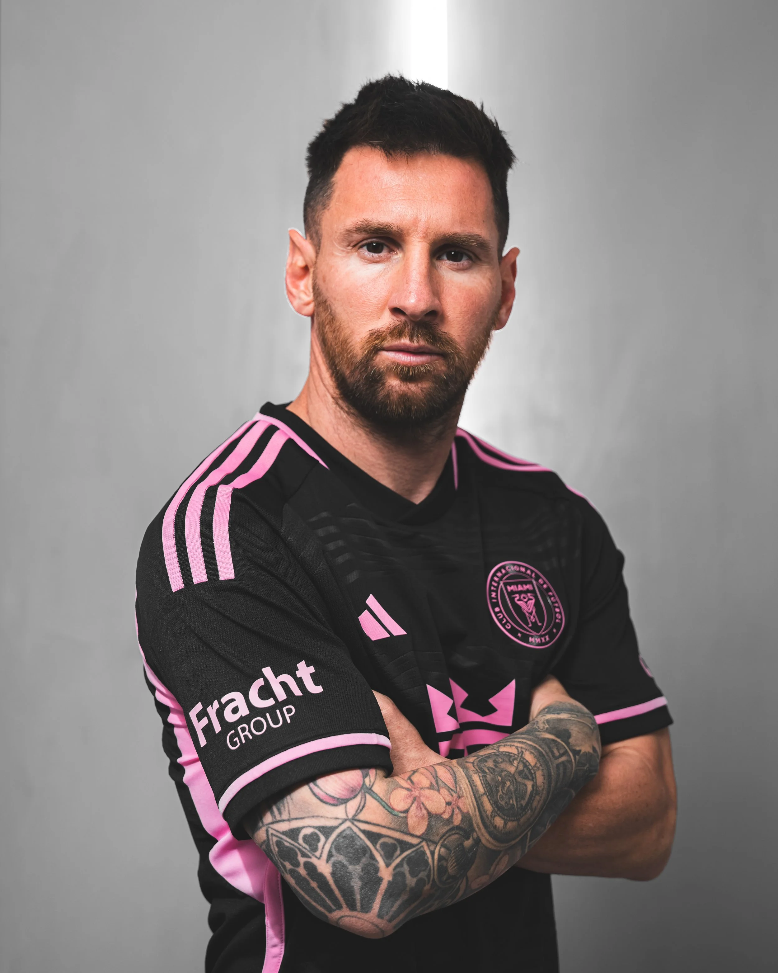 A man with short dark hair and tattoos on his forearm, wearing a black and pink sports jersey, standing against a plain gray background.
