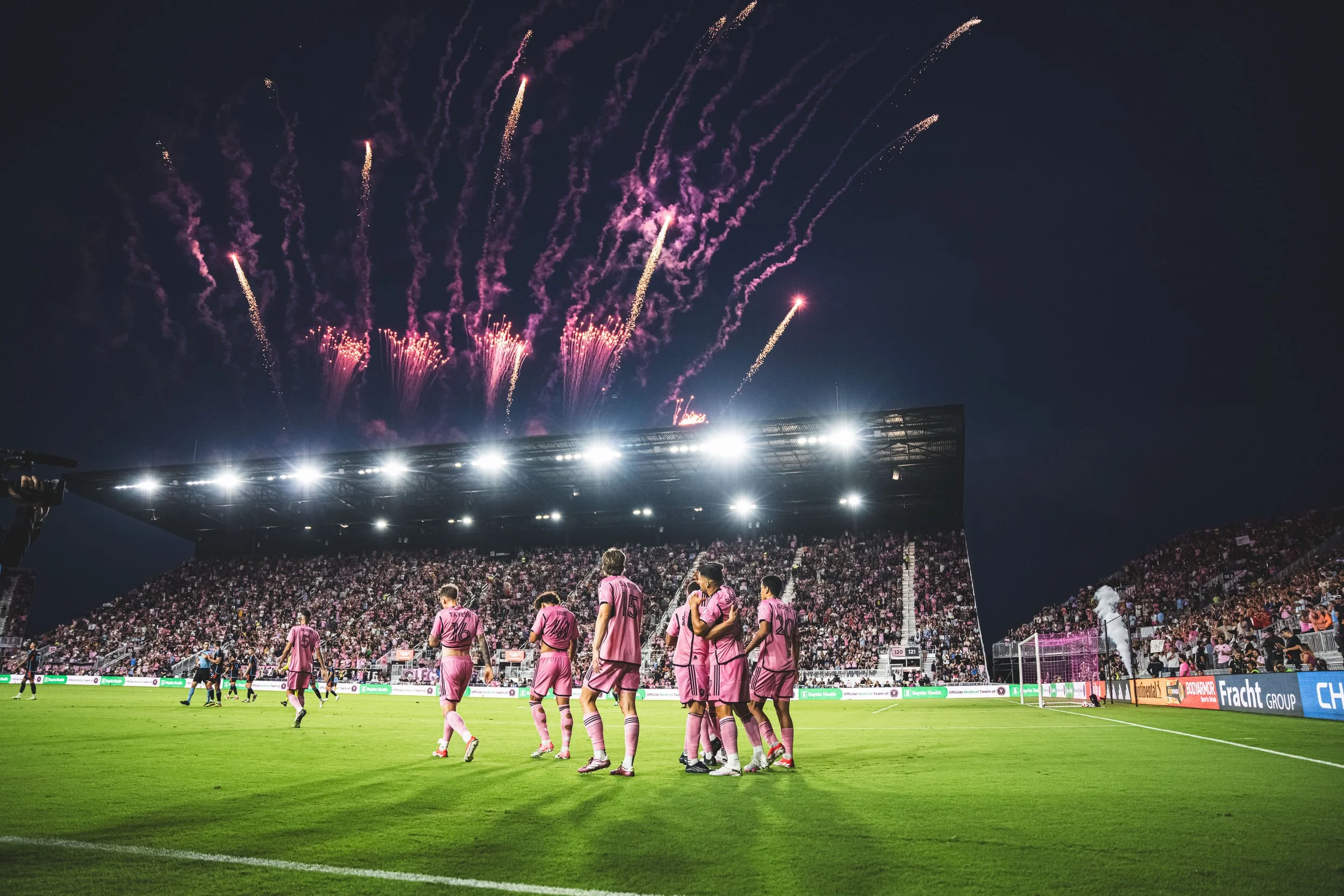 A soccer team in pink uniforms celebrating on the field with fireworks in the night sky above a large stadium filled with spectators.