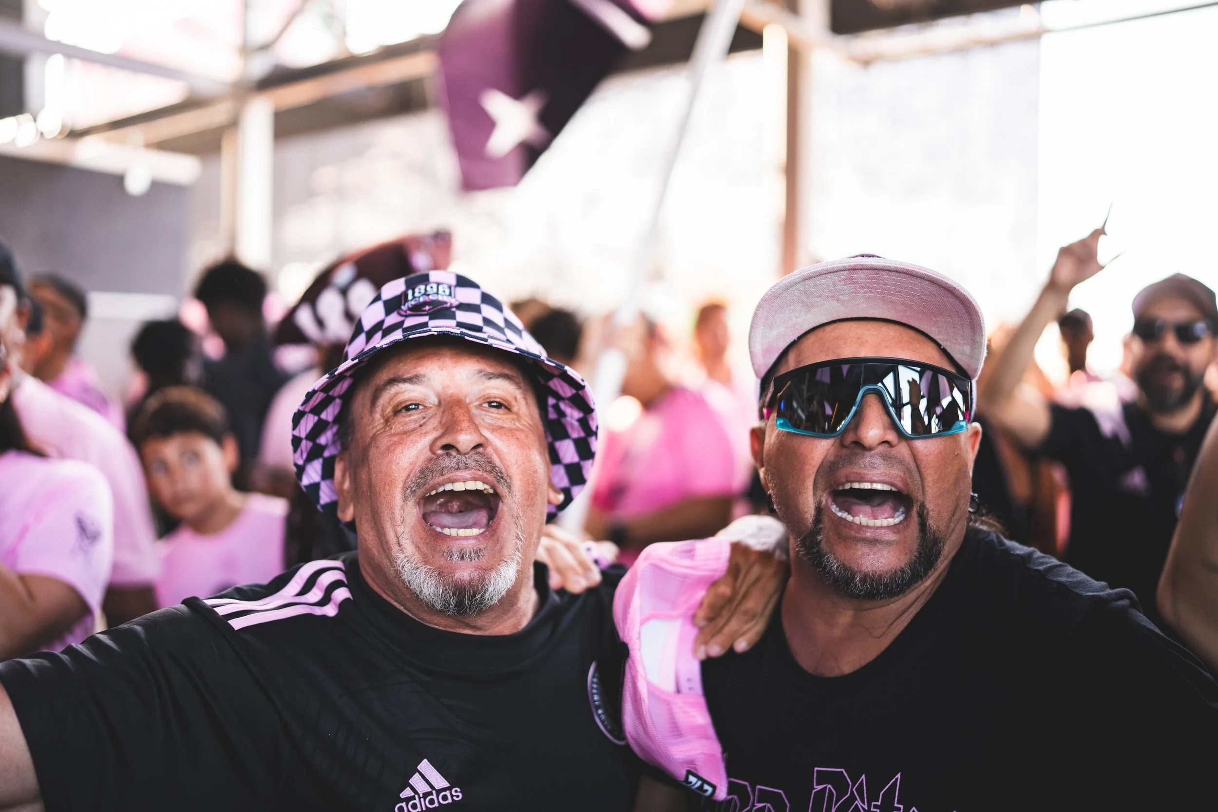 Two men celebrating at a crowded event, both with open mouths, one wearing a checkered hat and the other wearing sunglasses and a pink cap, amidst a group of people in pink shirts.