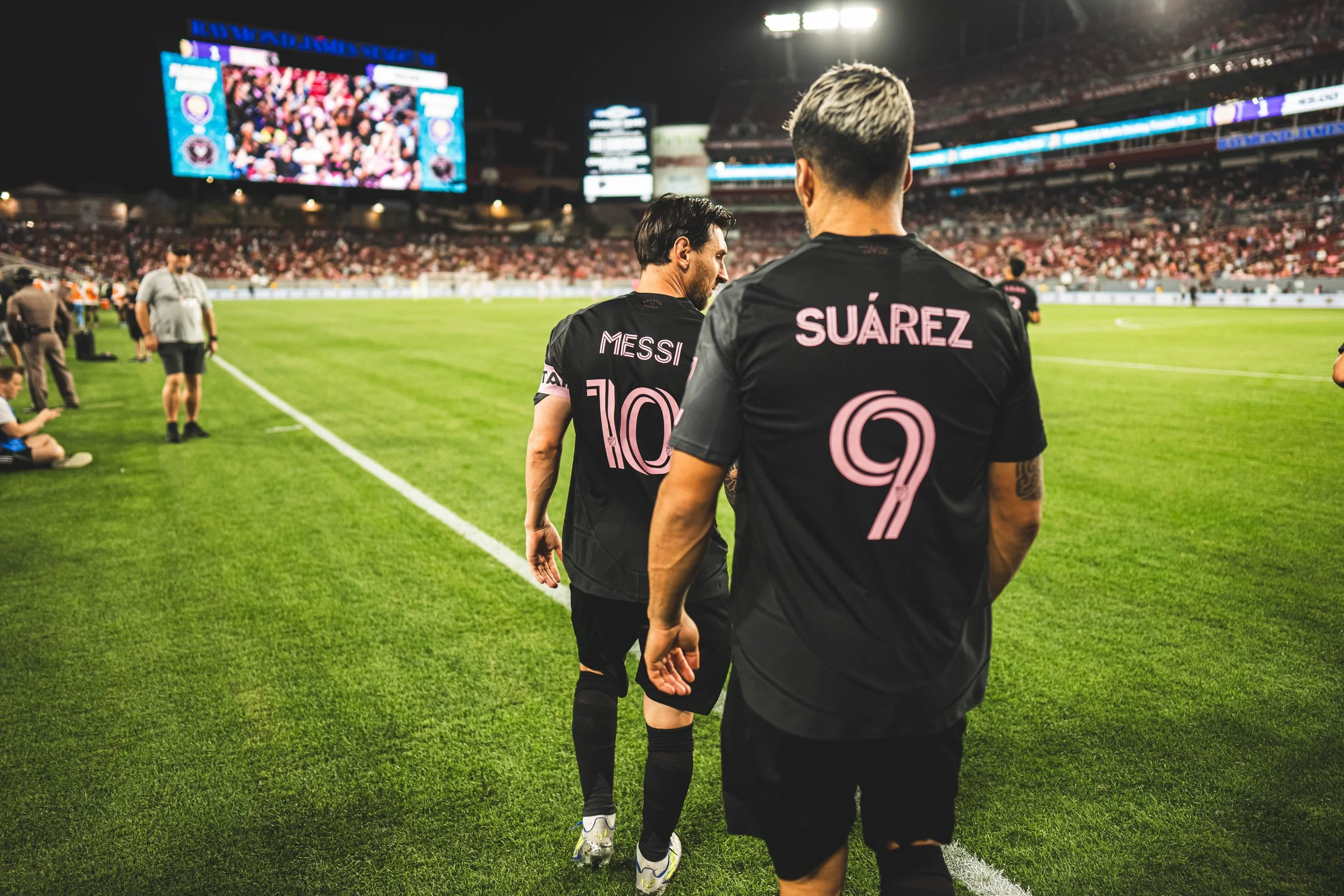 Soccer players Lionel Messi and Luis Suárez walking on the field during a nighttime match, with a large crowd and digital screens in the background.
