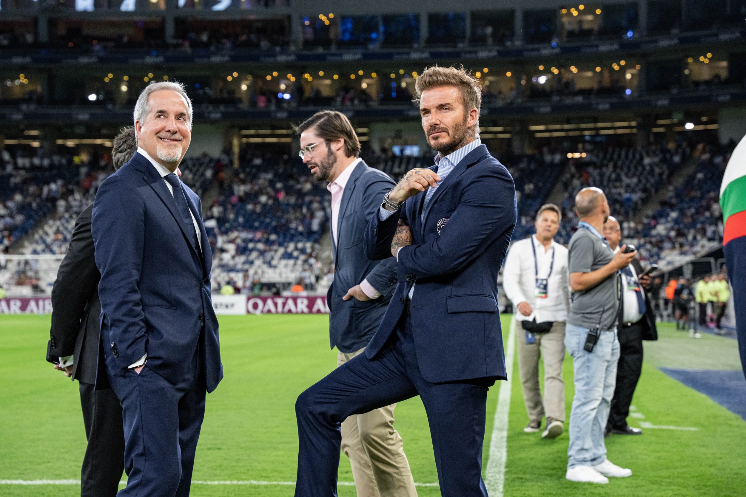 Three men in suits standing on a soccer field, with a crowd in the background, at a stadium during a sports event.