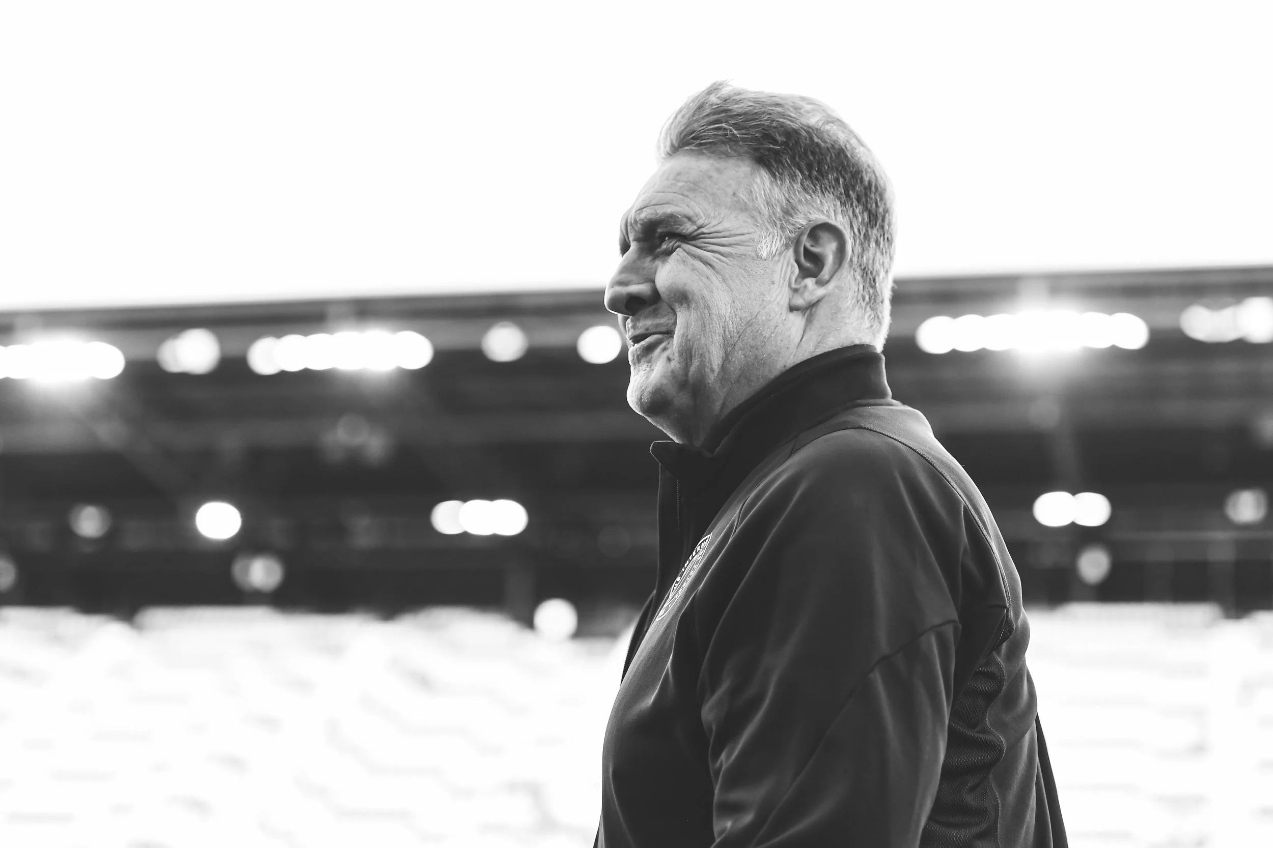 A black and white photo of a smiling man outdoors, wearing a sports jacket, with a stadium in the background.