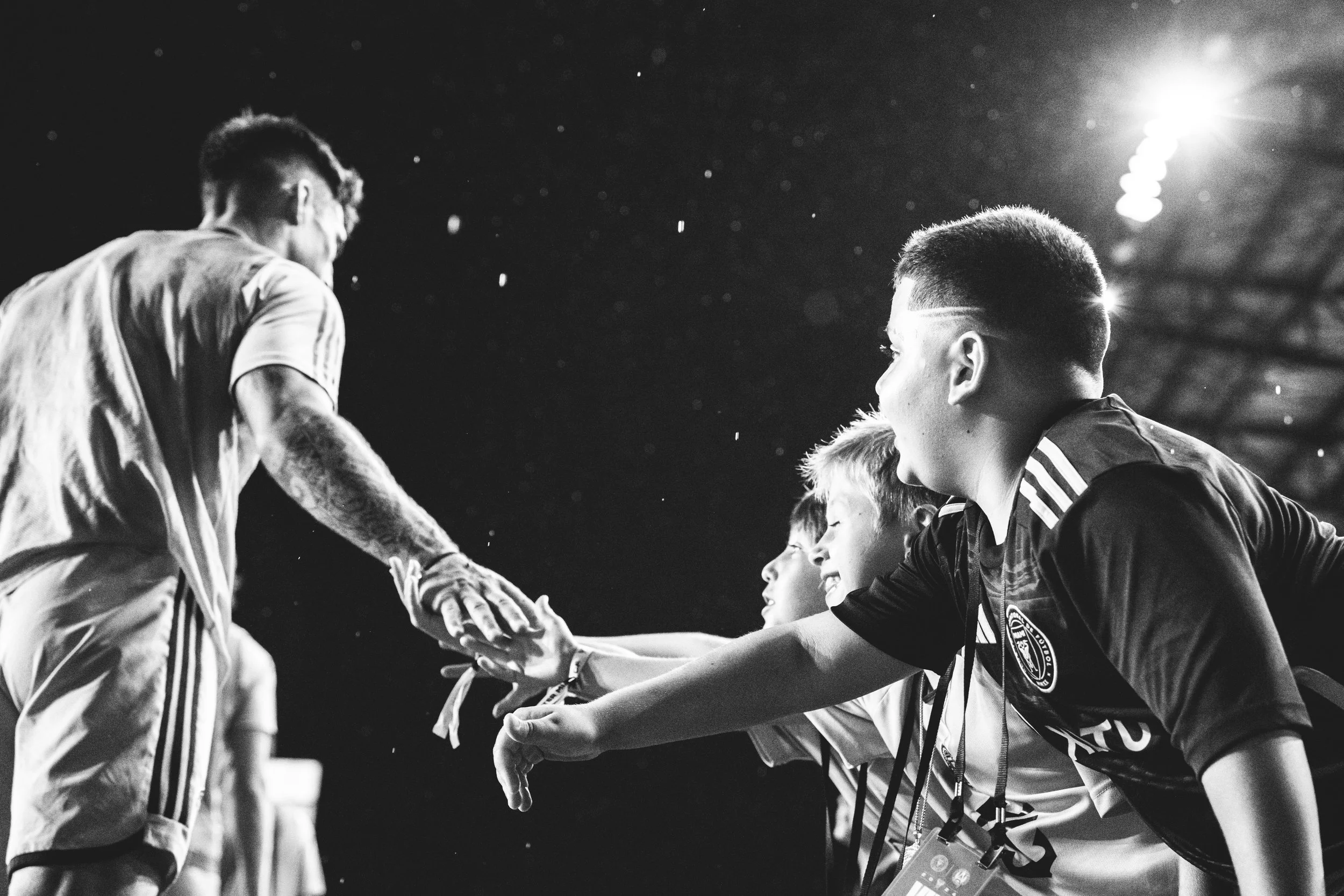 A group of children in sports jerseys reaching out to shake hands with a person in a sports uniform under stadium lighting.