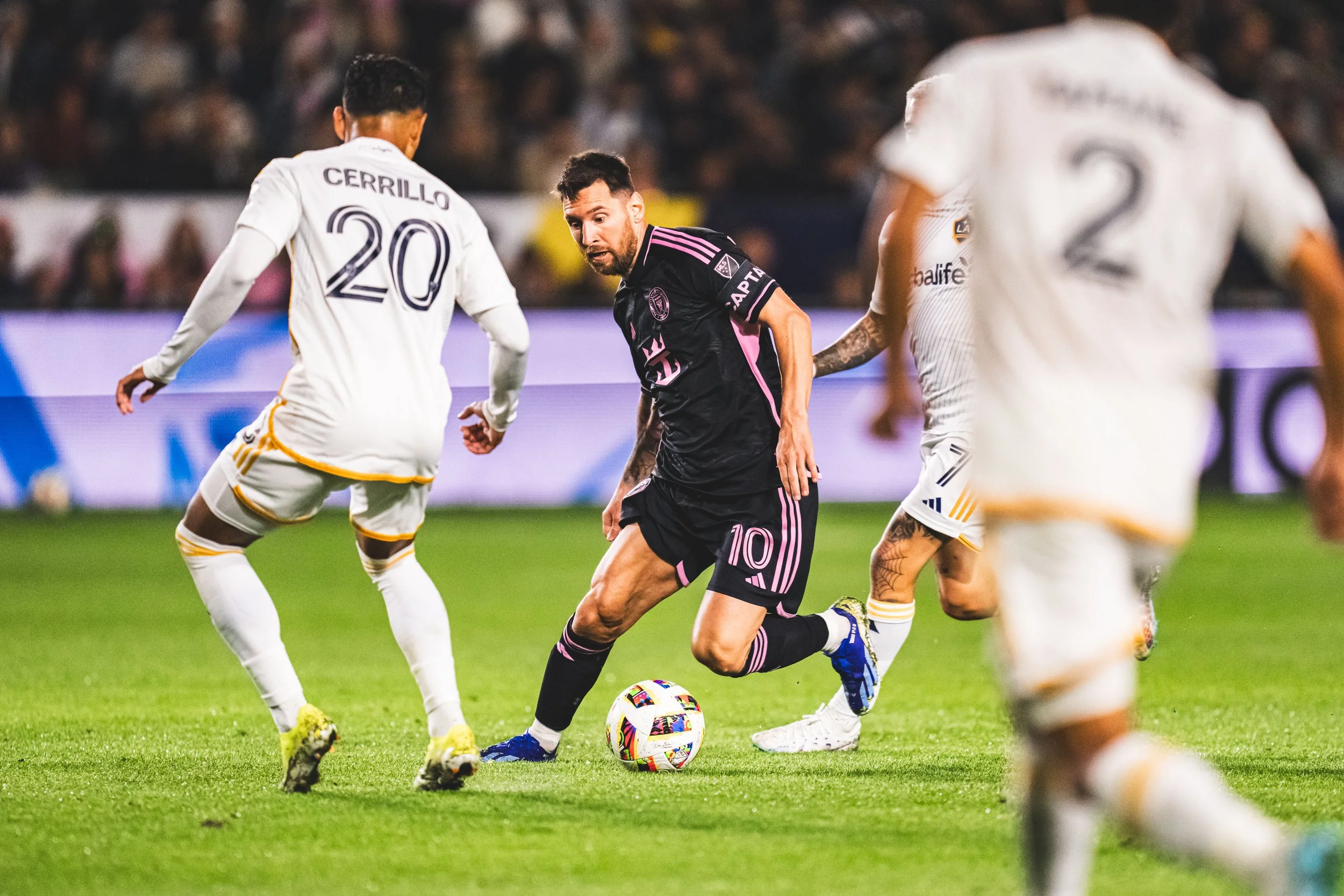 Soccer player with a beard in a black and pink kit controlling the ball during a match, surrounded by opponents in white kits on a green field with a crowd in the background.