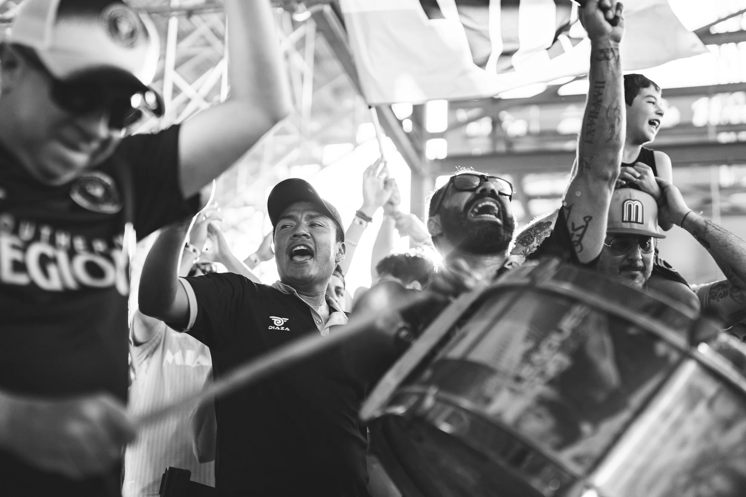 Group of people cheering and celebrating at a sports event, some holding a flag, with a focus on a man playing a drum, in a stadium.