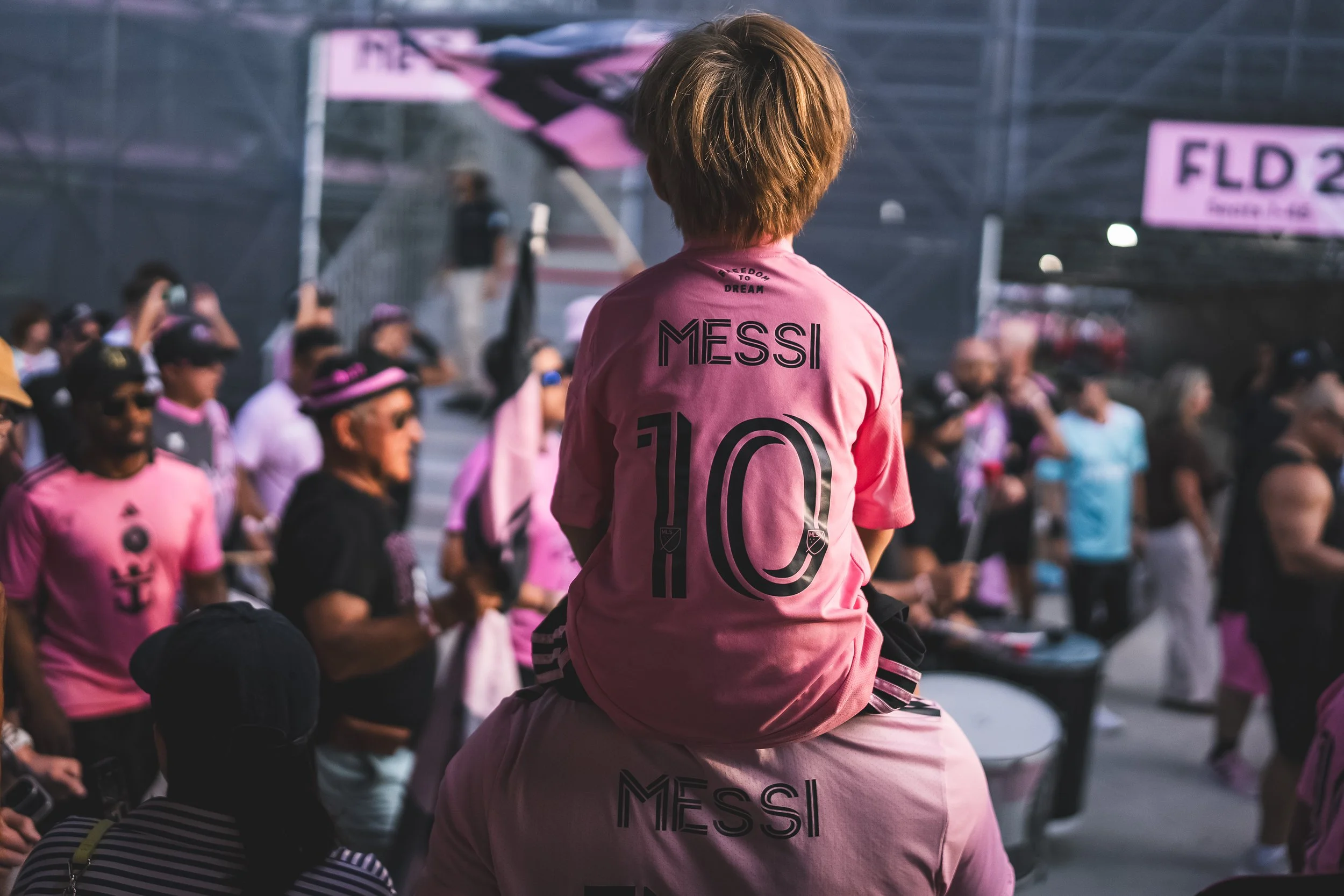 A young boy wearing a pink soccer jersey with 'MESSI 10' on the back sits on the shoulders of another person amid a crowd of people, many of whom are also wearing pink jerseys, at an outdoor event or rally.