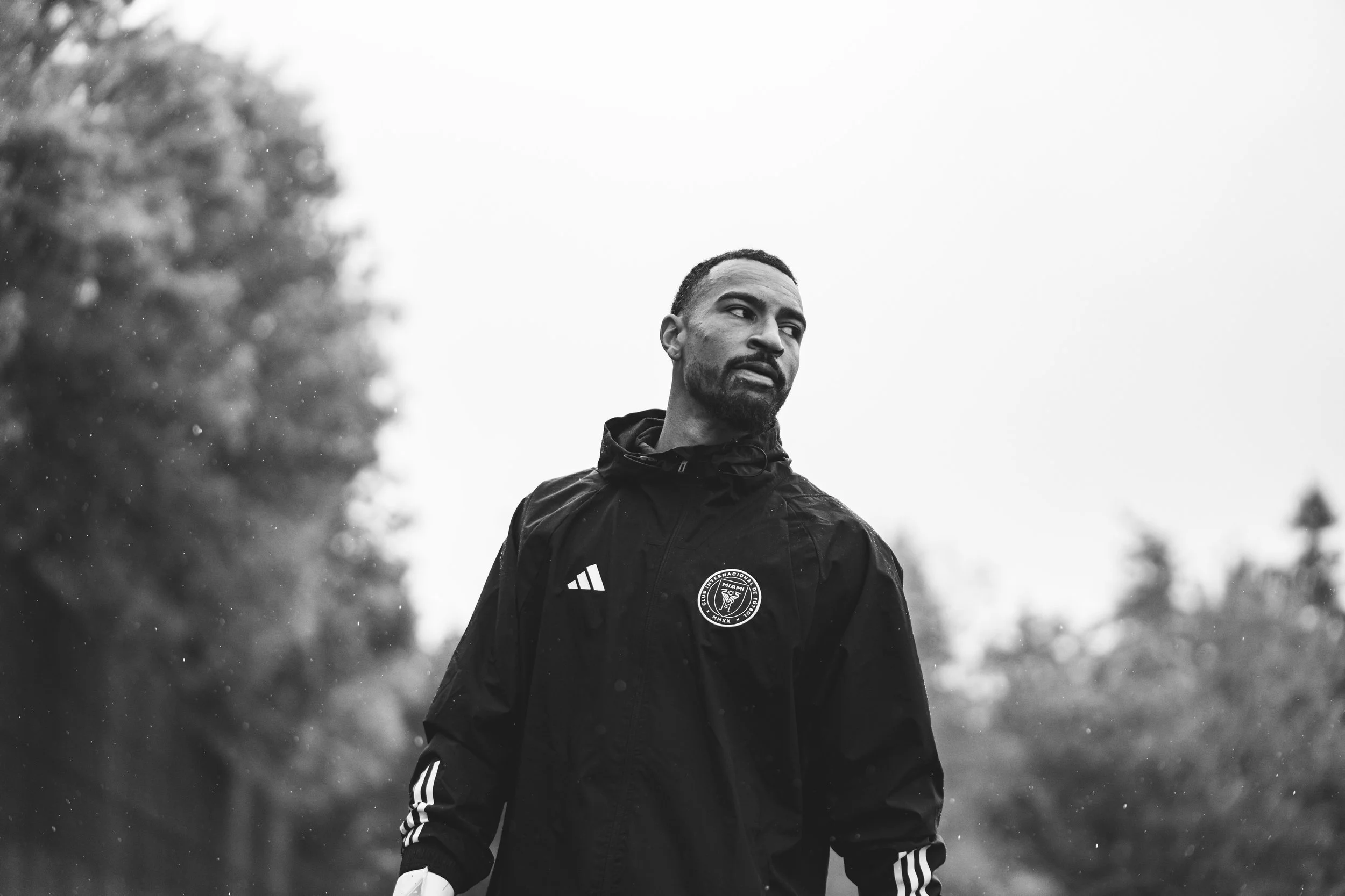 Black and white photograph of a man wearing a jacket with a sports team logo, outdoors with trees in the background, looking to the side.