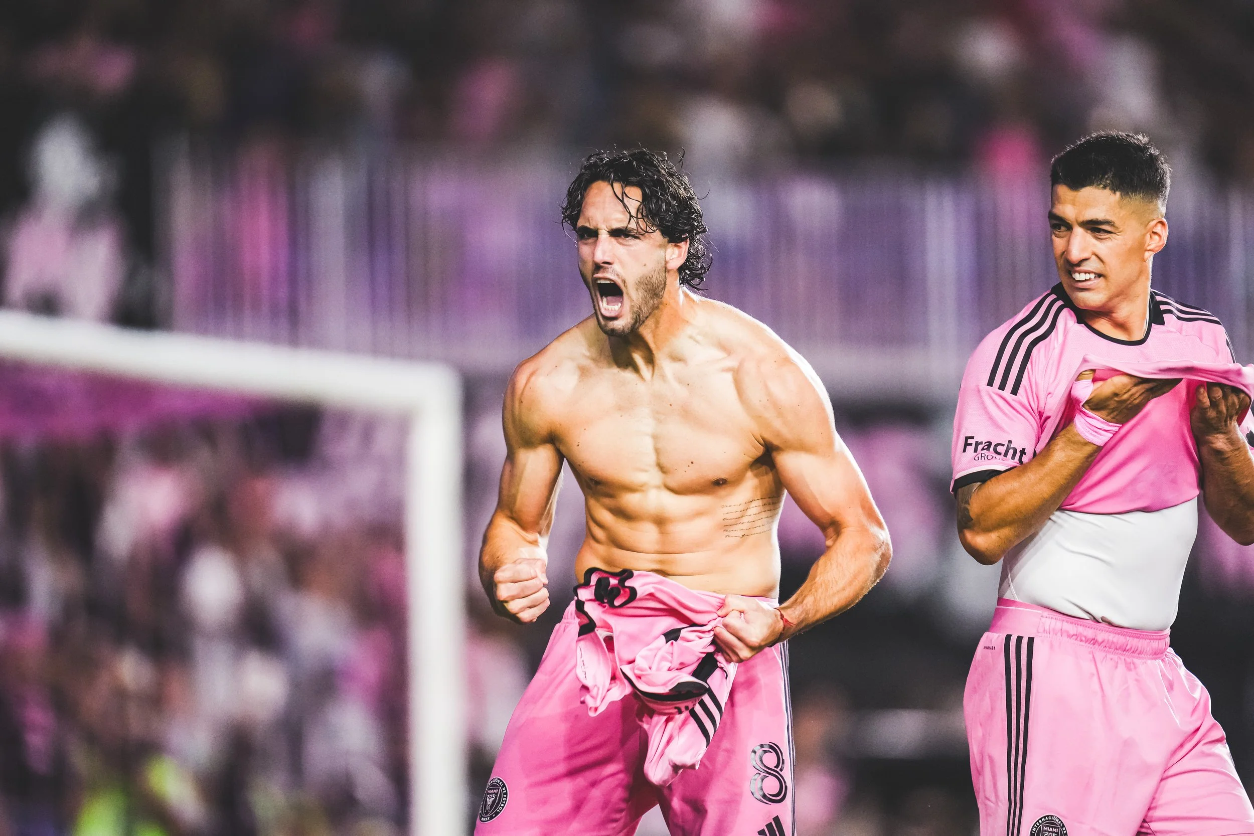 Two male soccer players, one shirtless and celebrating with a clenched fist and an open mouth, and the other holding his shirt to his chest, on a soccer field with a blurred background of spectators and a goalpost, wearing pink soccer uniforms.