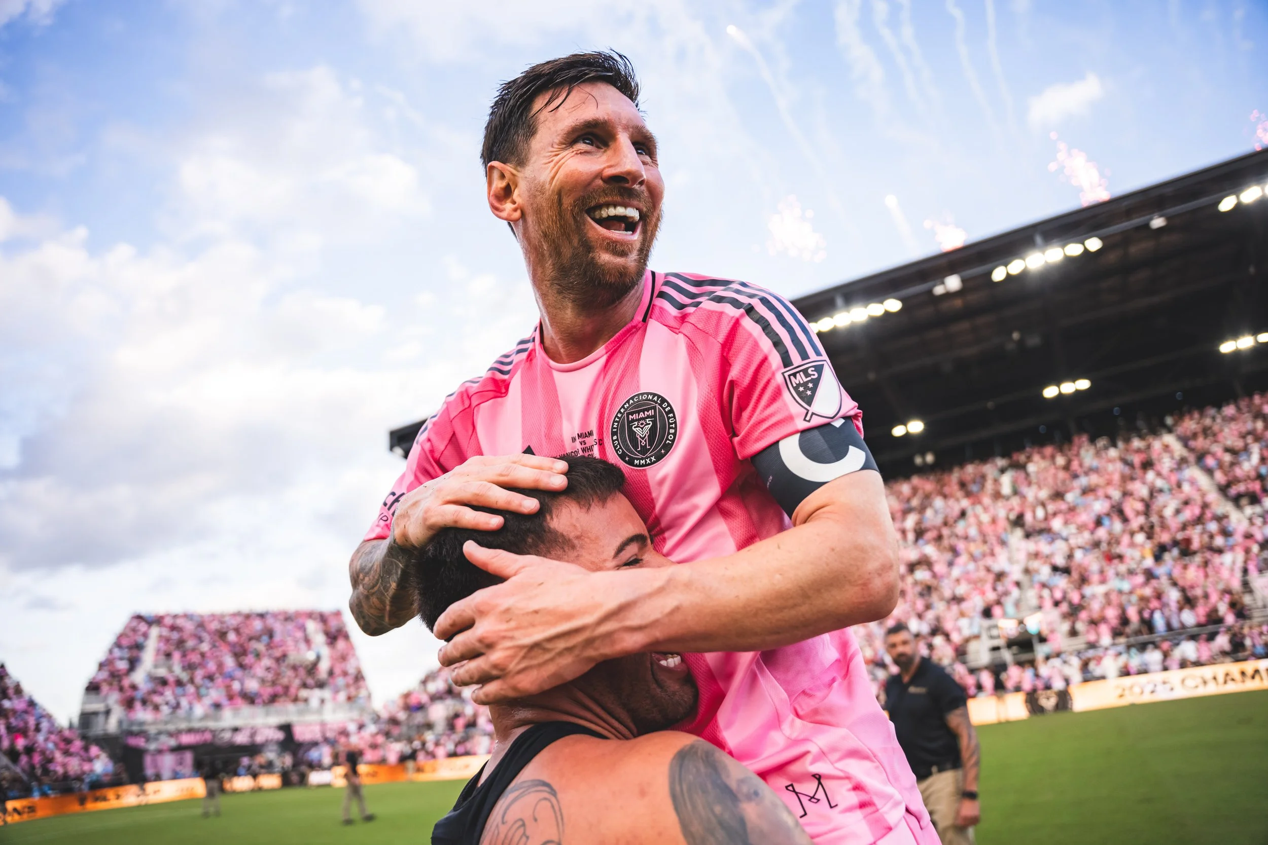 Two soccer players celebrating on the field, one in a pink uniform and the other in black, with a stadium full of pink-clad fans in the background.