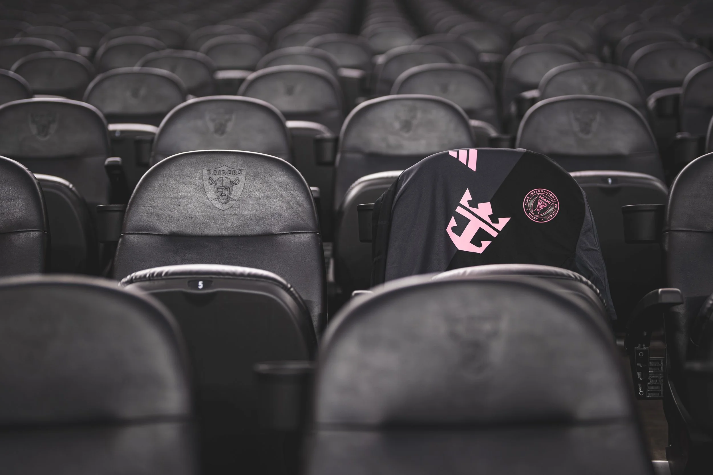 Empty black stadium seats with a Raiders logo on one, and a Miami FC backpack placed on another seat.