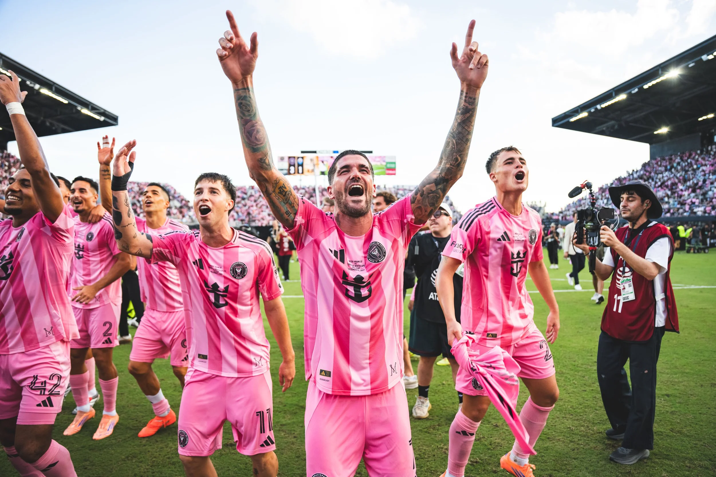 Group of soccer players in pink uniforms celebrating on the field with one player raising his arms.