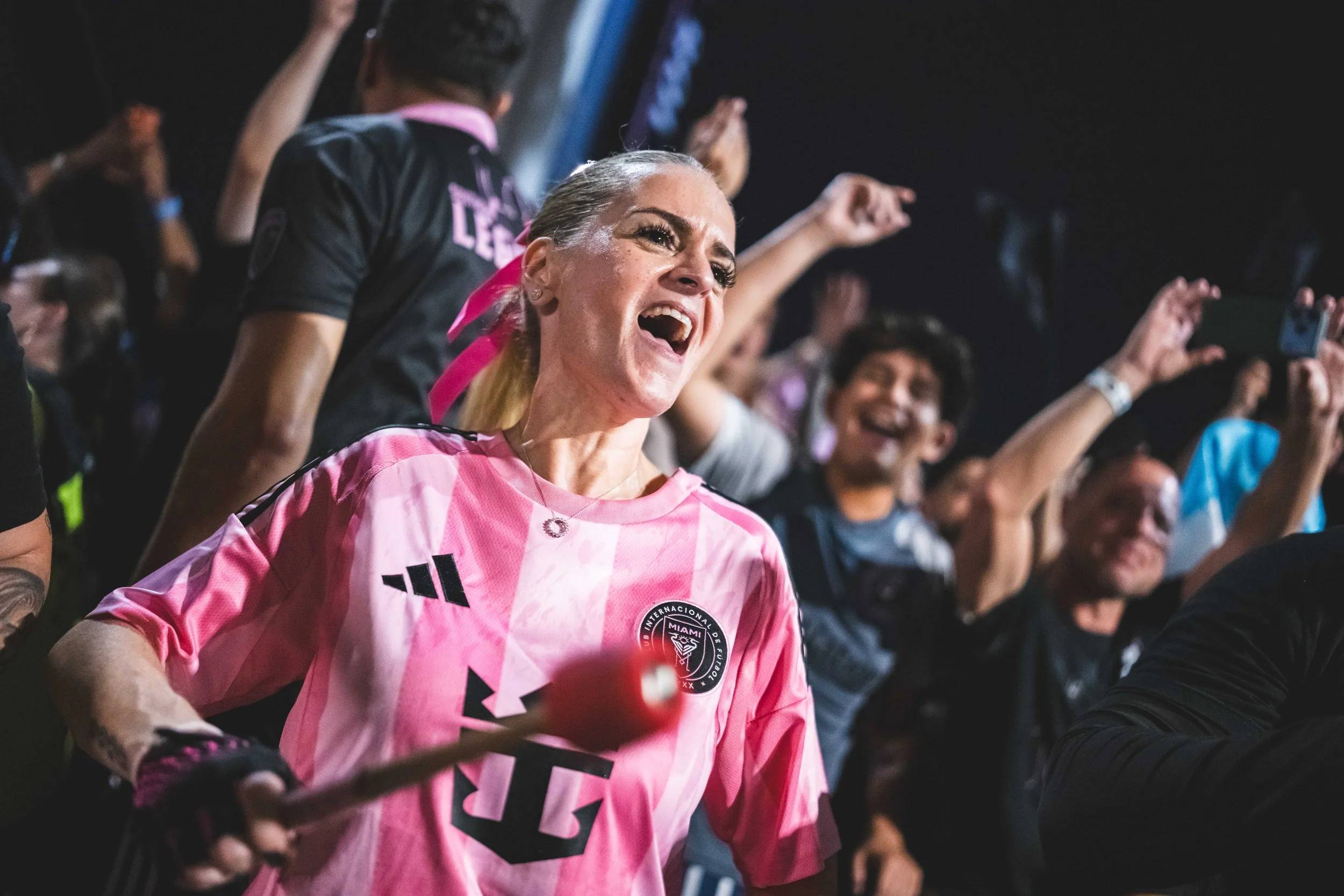 A woman in a pink sports jersey with a soccer team logo is celebrating enthusiastically among a crowd at a sports event, holding a mallet with a red rubber ball.