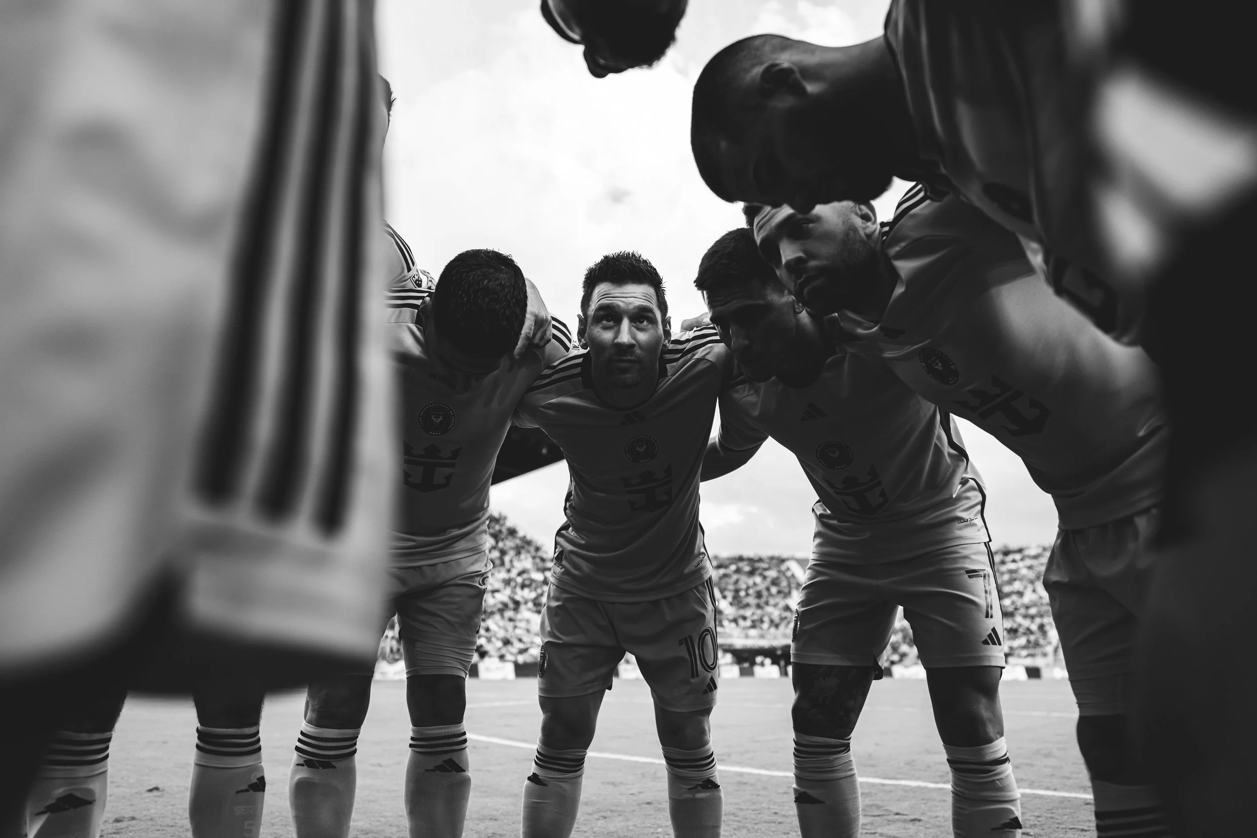 Black and white photo of a soccer team huddled in a circle on the field, with players leaning in, forming a close circle, facing inward. Sky and stadium in the background.
