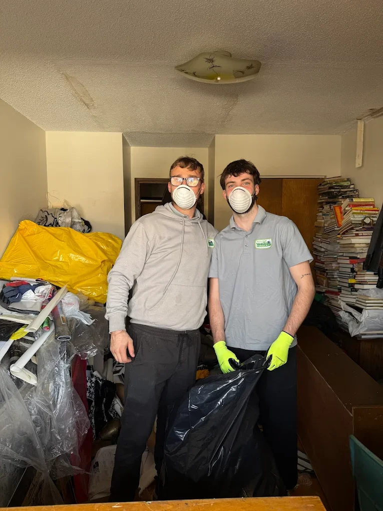 Two men wearing masks and gloves standing in a cluttered room with piles of books and trash, holding a black garbage bag.