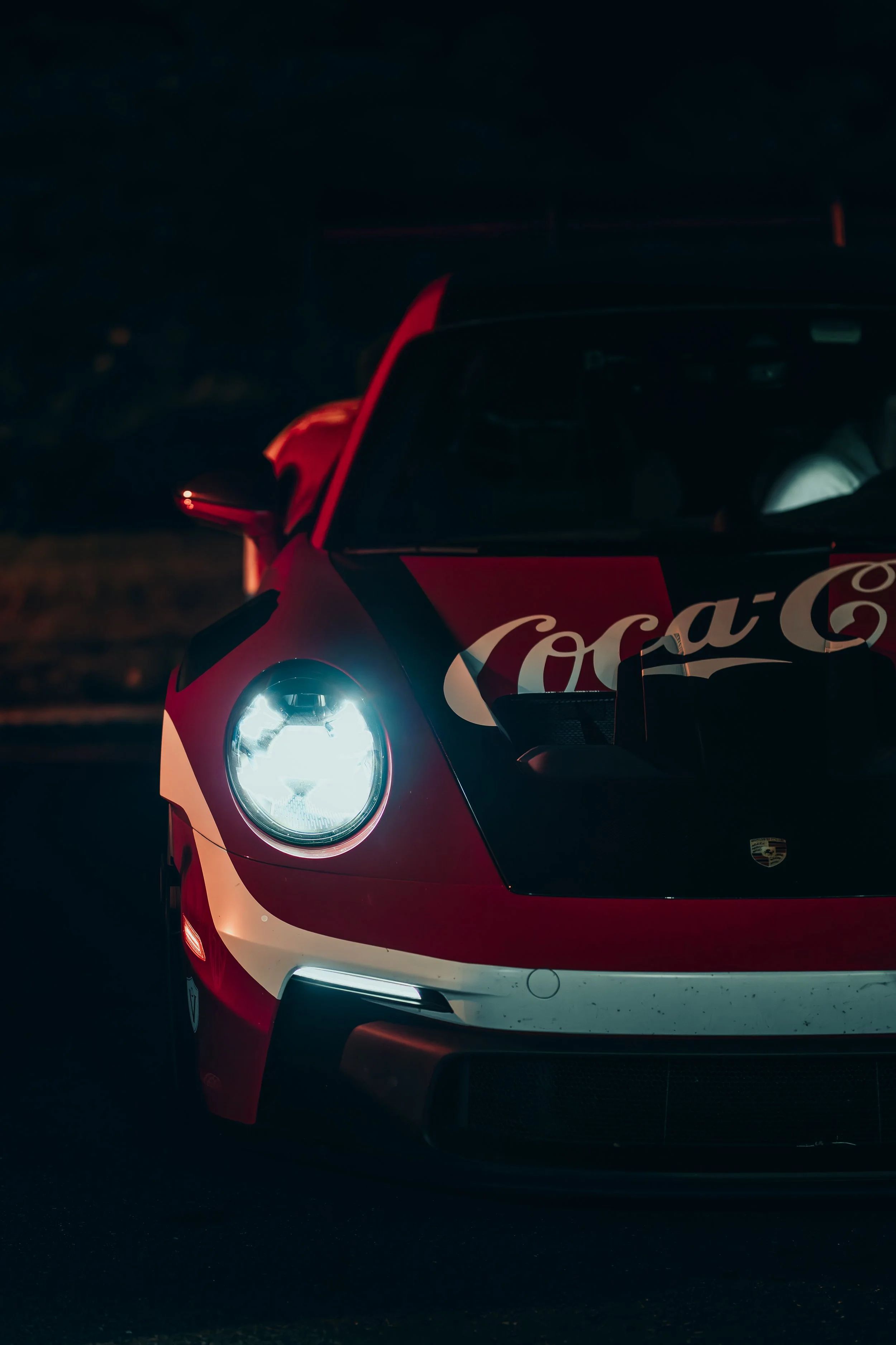 A Porsche race car with Coca-Cola branding, illuminated headlights, and a sleek red and white paint job, parked in the dark.