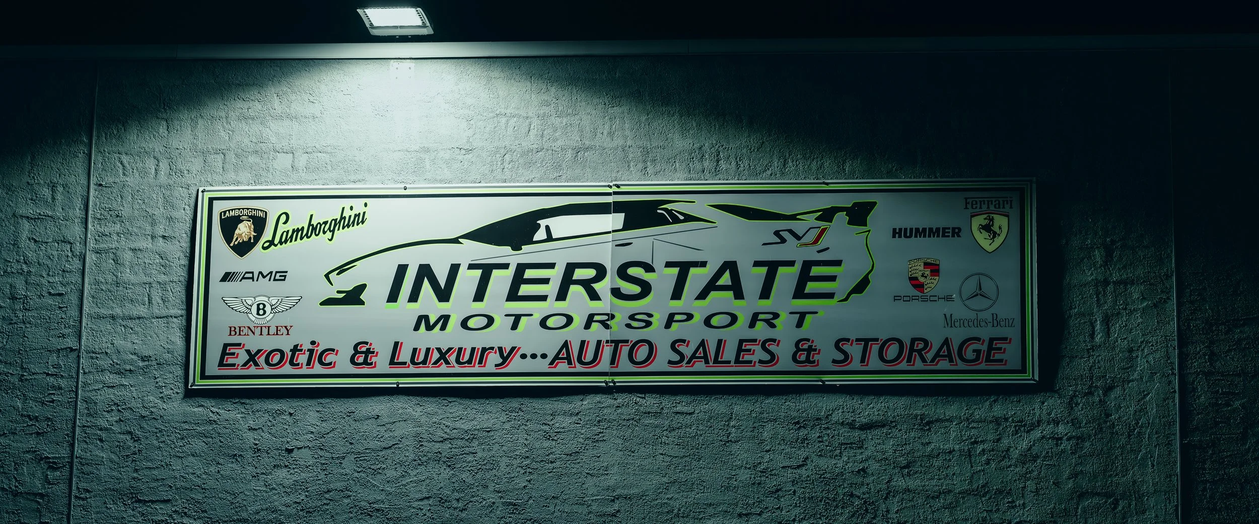 Sign for Interstate Motorsport advertising auto sales and storage, featuring luxury and exotic cars brands like Lamborghini, Ferrari, Hummer, Porsche, Mercedes-Benz, and Bentley, mounted on a textured wall with overhead lighting.