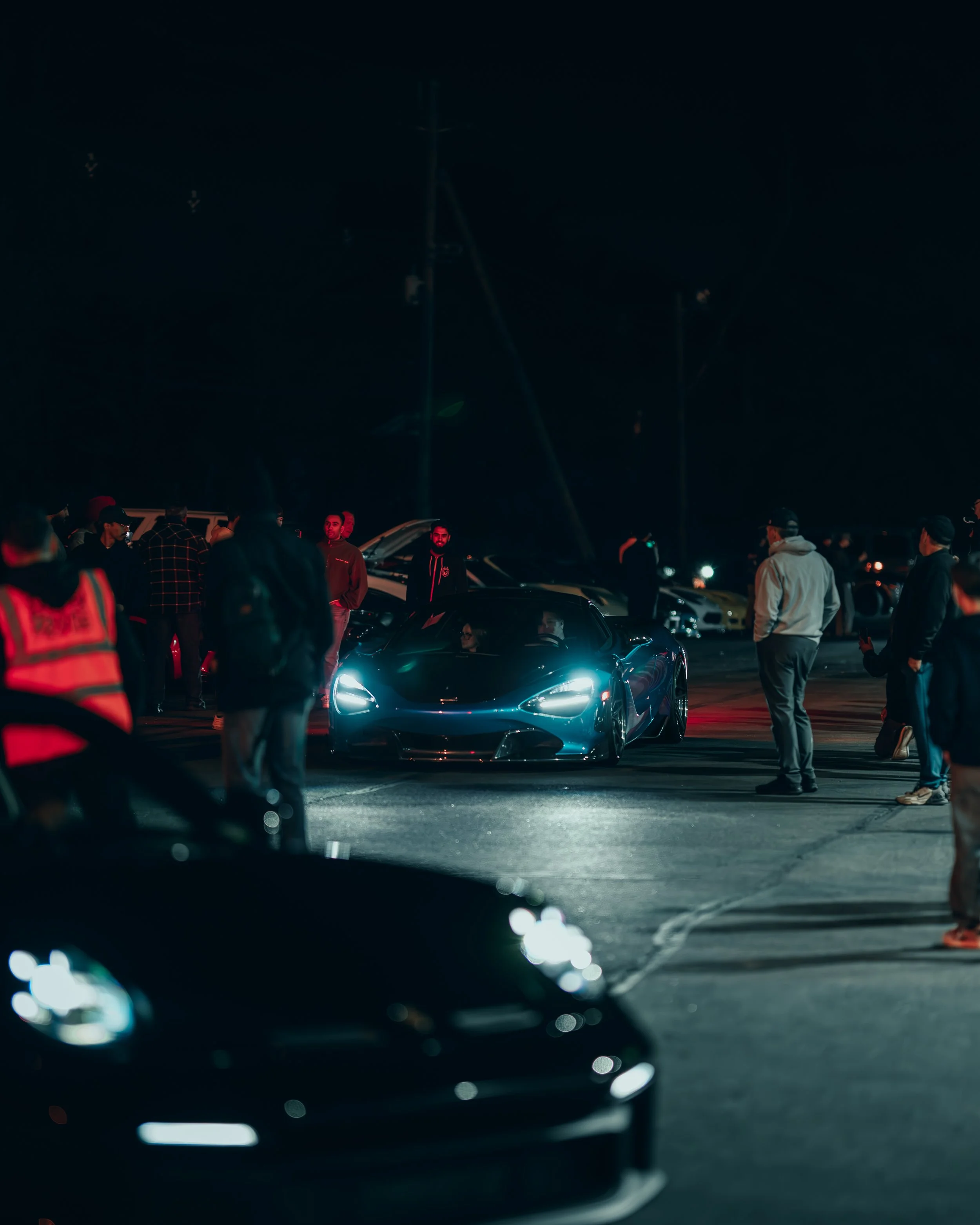 Nighttime scene at a car meet with people gathered around a blue sports car with its headlights on, surrounded by other cars and spectators.