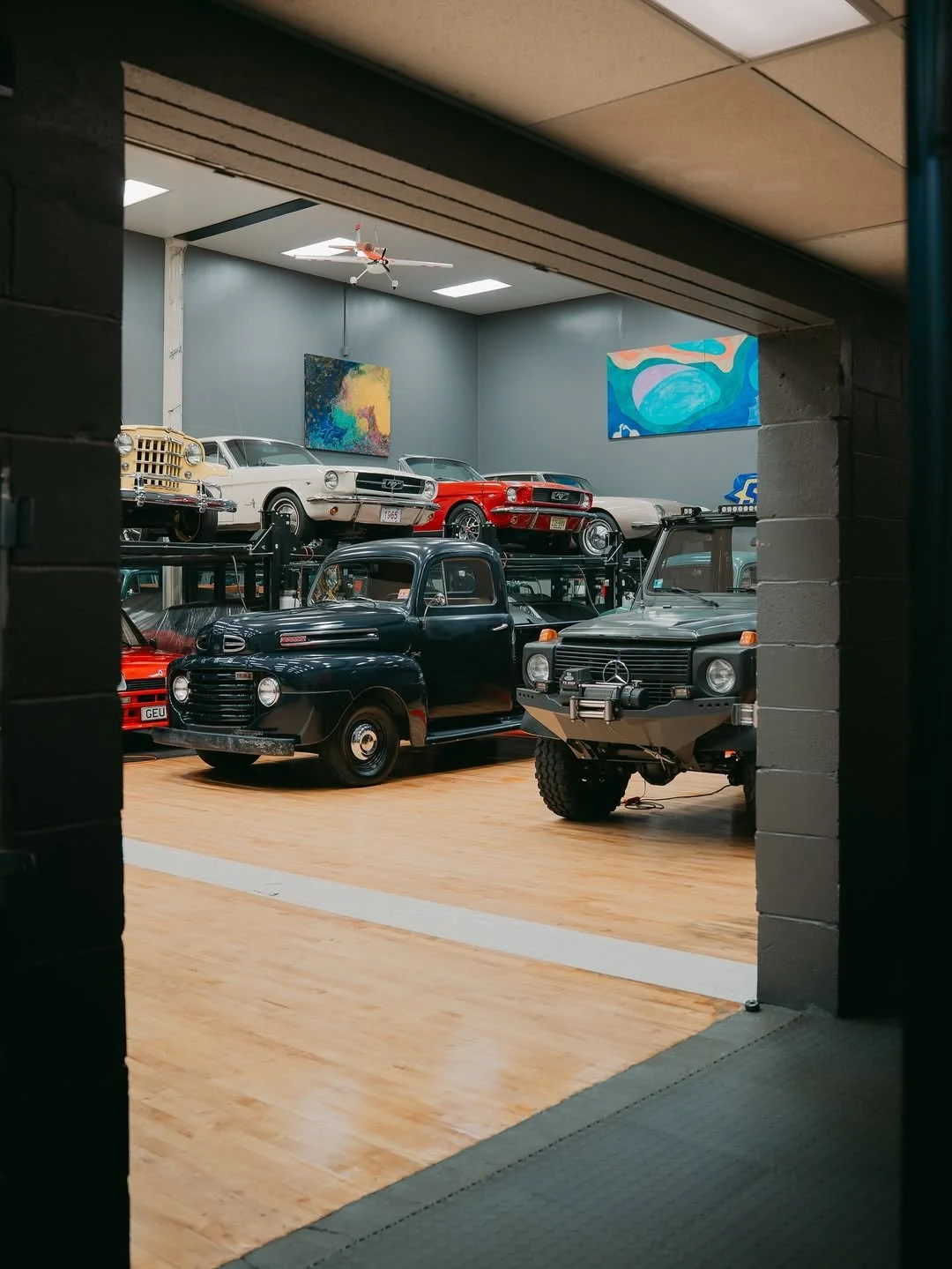 View of a vintage car museum with various classic cars displayed on a wooden floor, including a black truck, a light-colored truck, and a red sports car, with artworks hanging on the blue wall and a miniature airplane hanging from the ceiling.