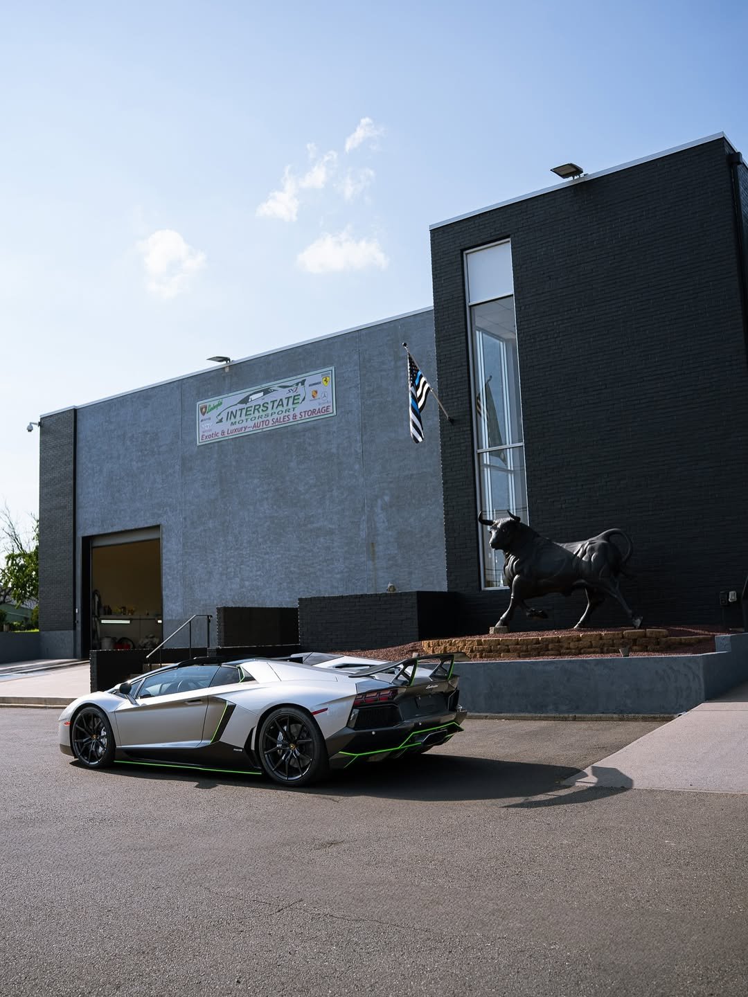 A luxury silver Lamborghini sports car parked outside a modern building with a black and gray exterior, a statue of a bull, and an American flag on a pole, under a blue sky with scattered clouds.