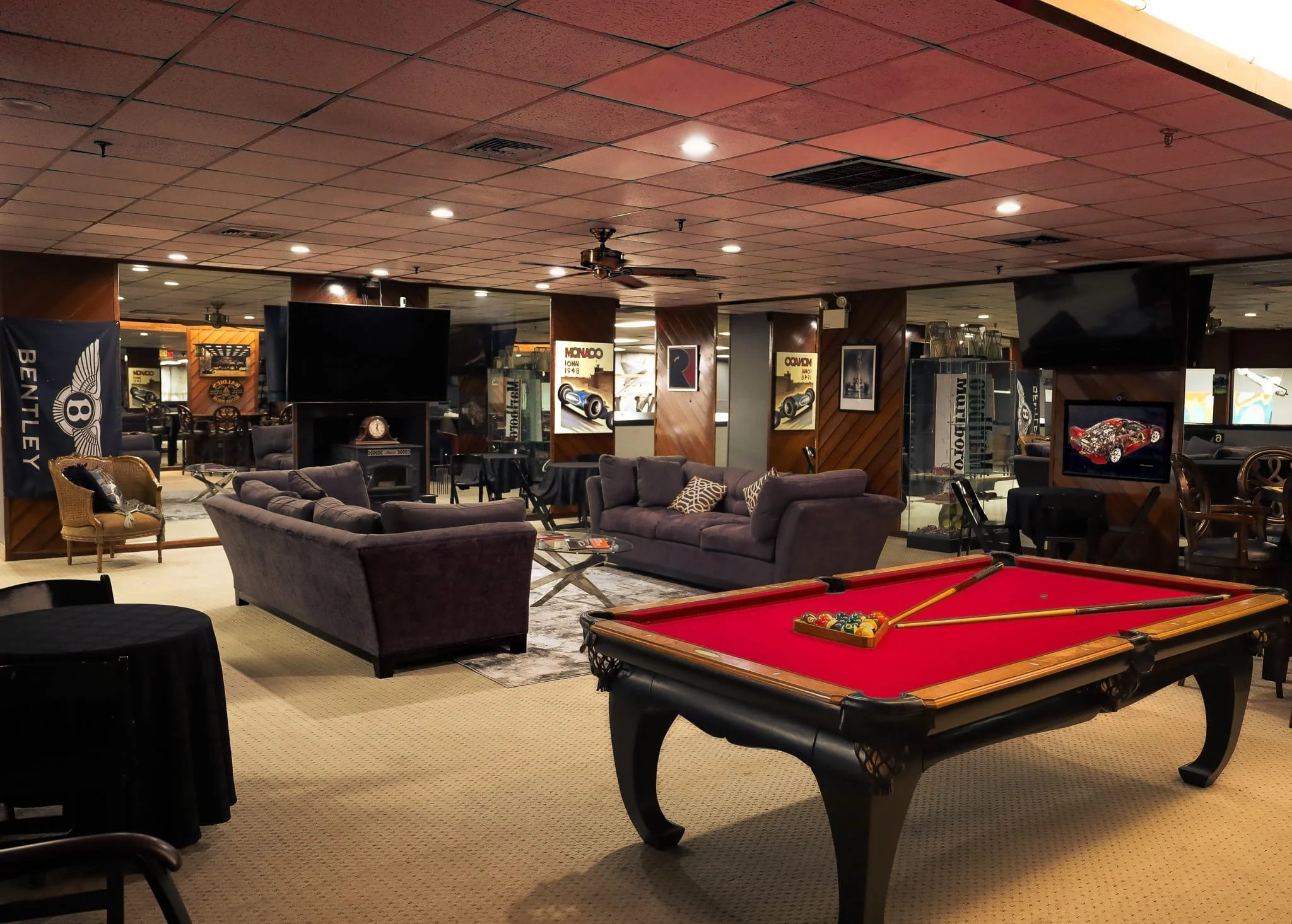 Indoor lounge area with purple sofas, a pool table with red felt, wall-mounted televisions, framed posters, and vintage car memorabilia.