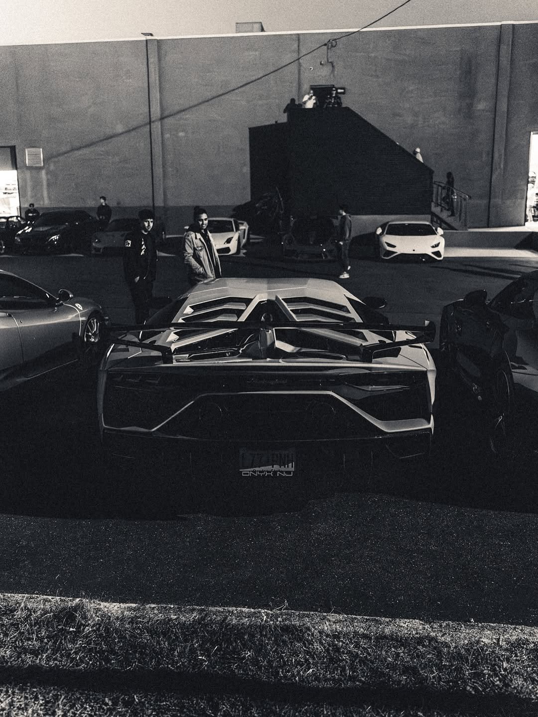 A black and white photo of a parking lot with luxury cars, including a Lamborghini, with three people standing near the cars and some others in the background.