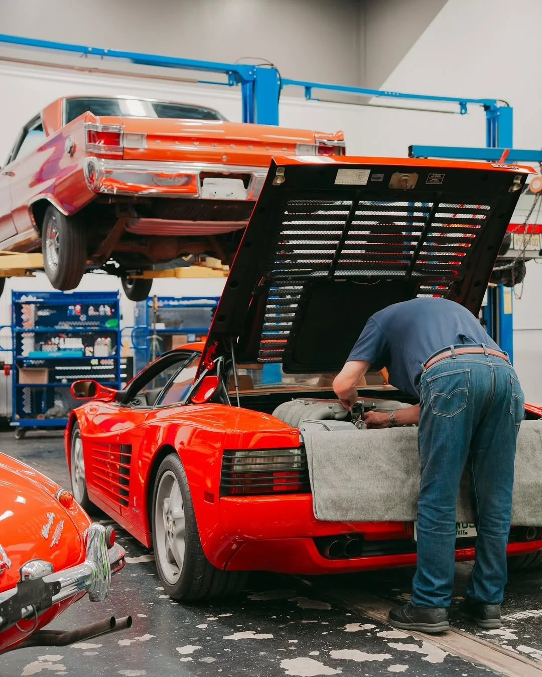 A mechanic working on a red sports car with its hood open in an auto repair shop. In the background, a red vintage car is lifted on a hydraulic lift.