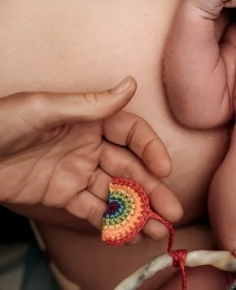 A person's hand holding a small, colorful crochet rainbow near their breast.