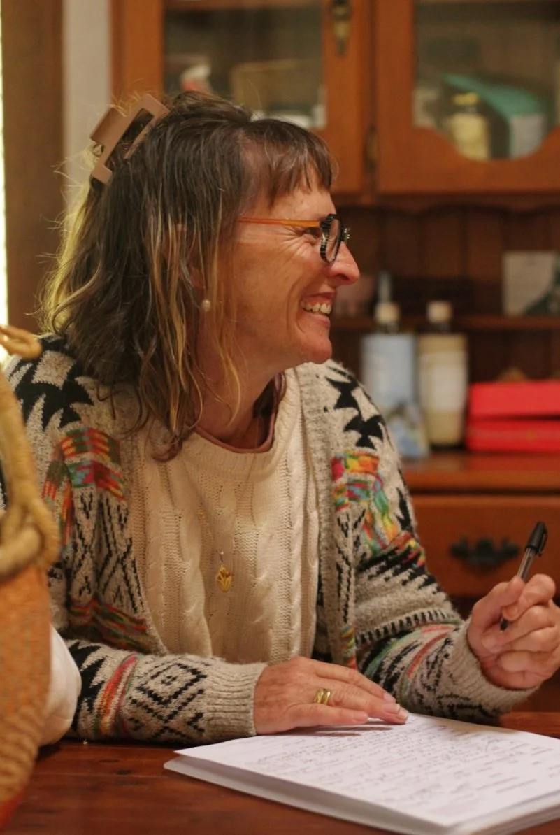 A woman with glasses and a patterned sweater, smiling and sitting at a wooden table with papers in front of her, in a warm, cozy kitchen.