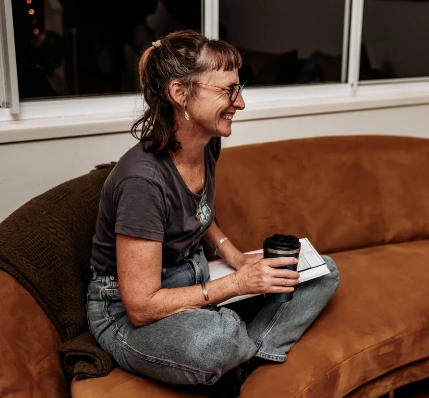 A woman with glasses, earrings, and short brown hair tied back, smiling and sitting cross-legged on a brown couch, holding a coffee cup and a notebook, in a living room.