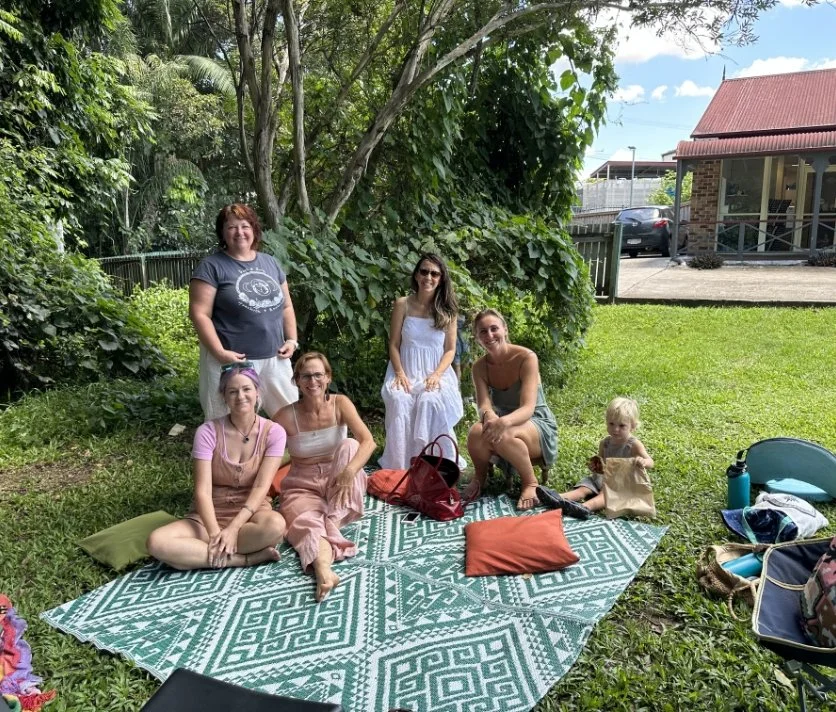 Six women and a child sitting and standing on a patterned blanket in a grassy backyard with green trees and a house in the background.