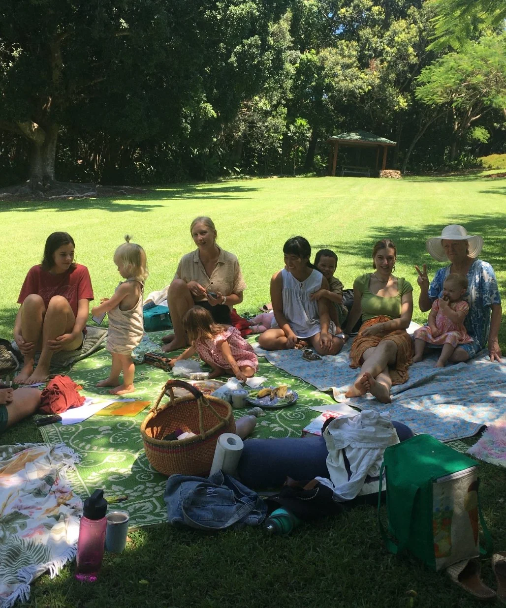 A group of people, including children and adults, having a picnic on a grassy area with trees in the background. They are sitting on blankets with various picnic items around them.