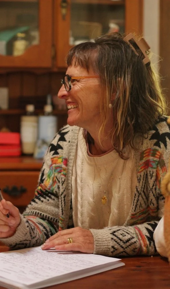 A middle-aged woman with glasses, shoulder-length wavy hair, smiling and sitting at a wooden table. She is wearing a cream sweater with black patterns and colorful details, a necklace, and a gold ring. Behind her is a wooden cabinet with various bottles and items.