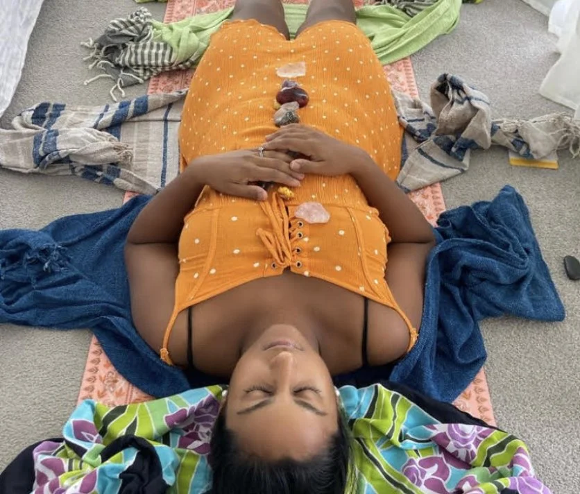 A person lying on their back on a mat with their eyes closed, wearing an orange top and yellow skirt. They have various crystals placed in a line on their torso and are surrounded by colorful cloths and towels.