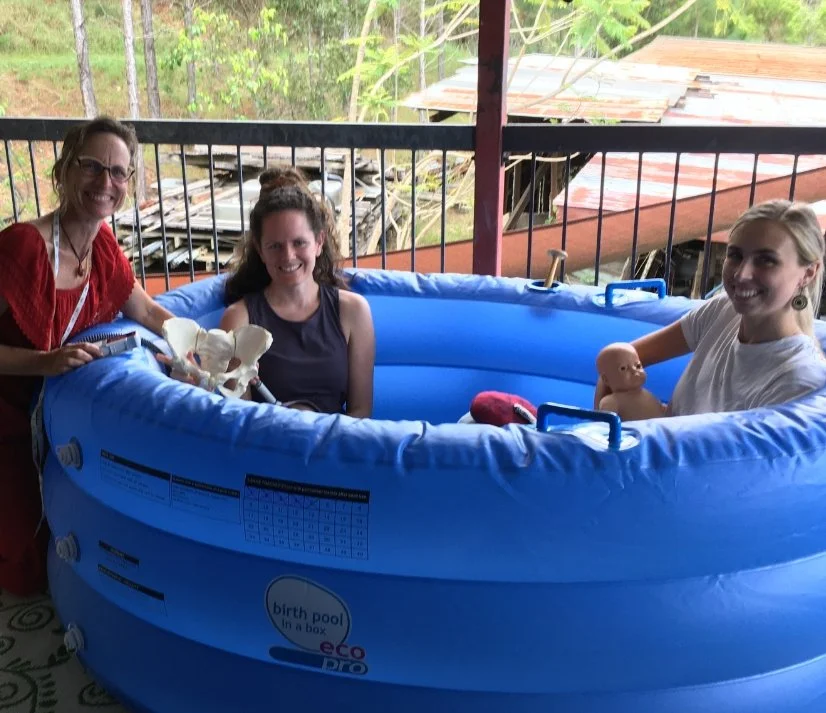 Three women sitting in a blue inflatable birth pool on a porch, holding a mannequin doll and a baby doll, with a skeleton prop. One woman is standing outside the pool, while two are sitting inside holding the dolls, and the porch has a metal railing with trees and rooftops visible in the background.