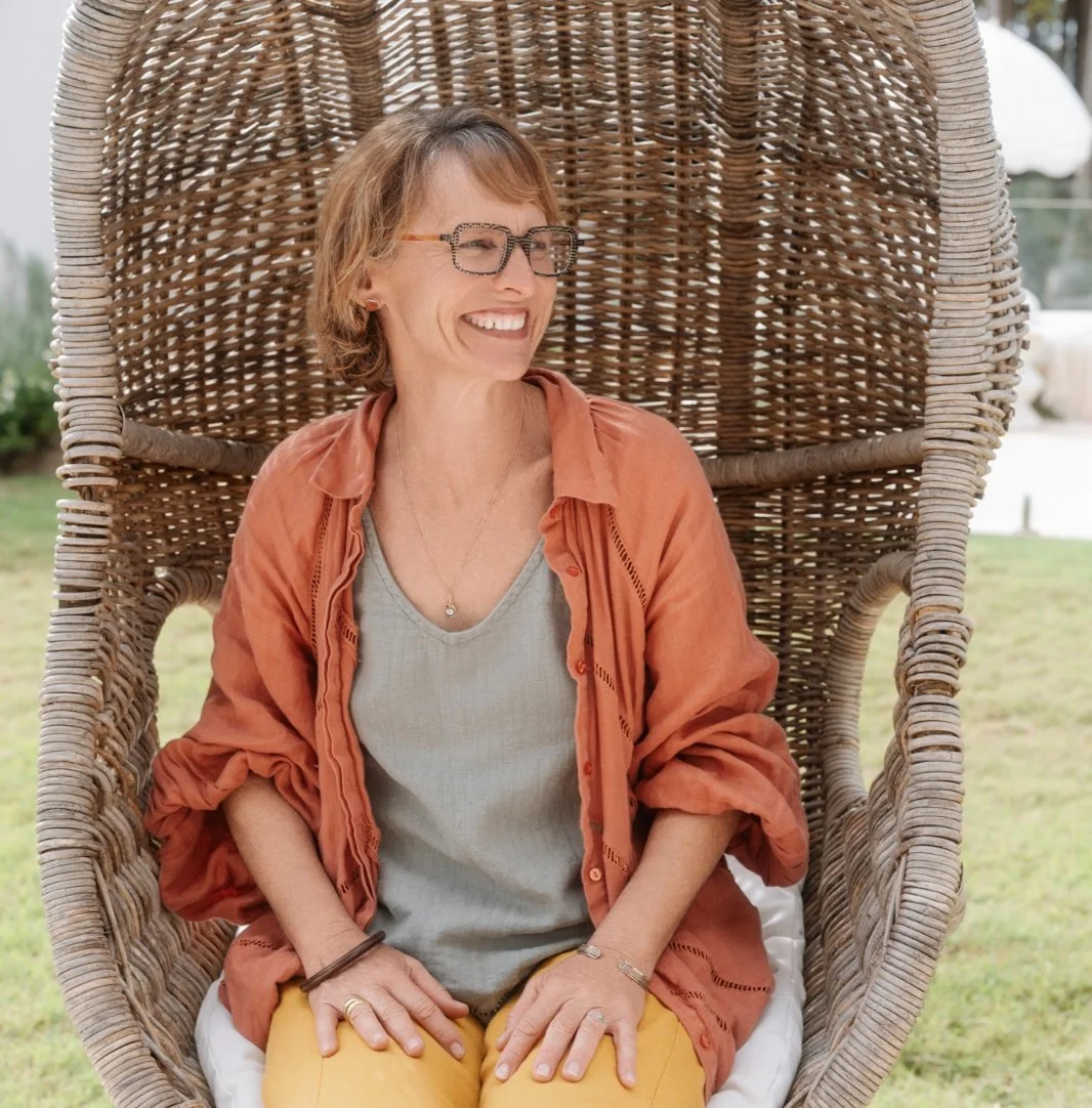 A woman with short reddish hair and glasses, smiling while sitting in a large wicker chair outdoors.