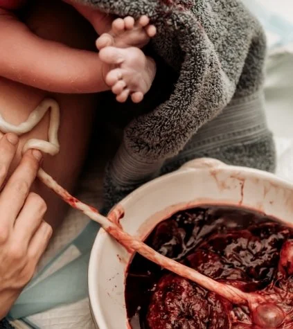 Close-up of a newborn baby suckling at the mother's breast with a cord seen in the scene, and a bowl of blood and entrails nearby.