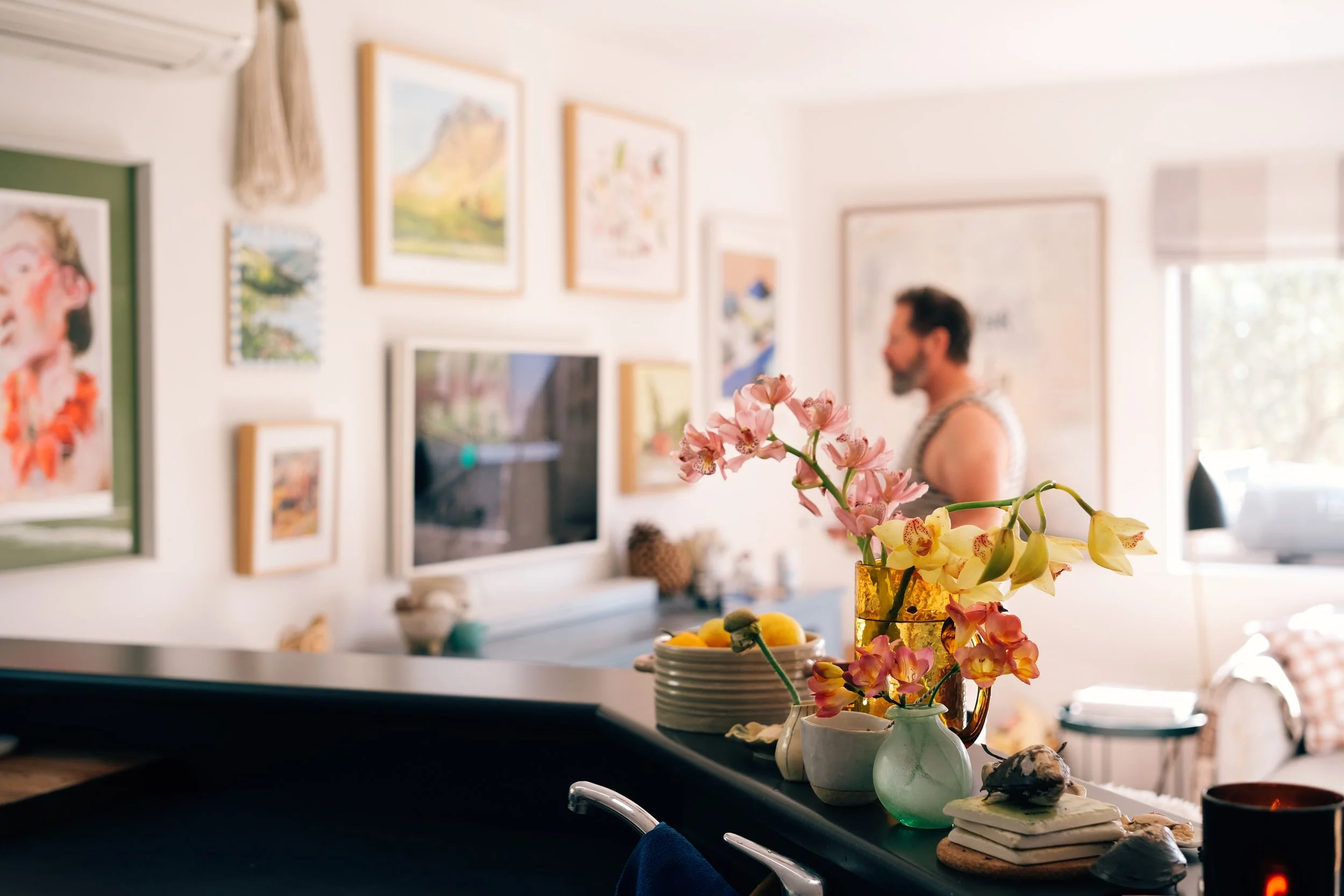 Close-up of pink and yellow flowers in vases on a kitchen counter with a blurred man in a sleeveless shirt in the background, inside a well-lit living room decorated with framed artwork.