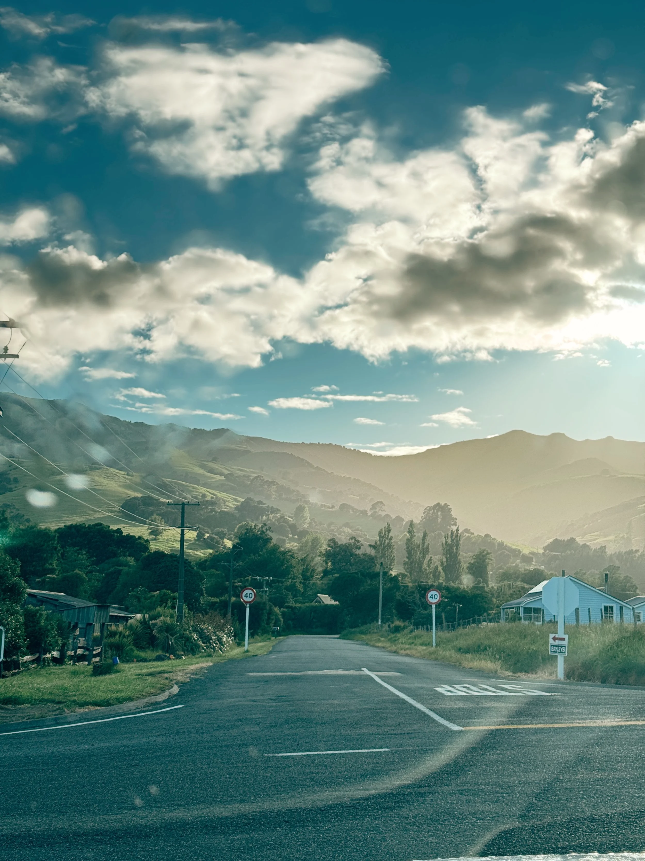 A rural road leading towards green hills with a mountain range in the background, under a partly cloudy sky with the sun shining.