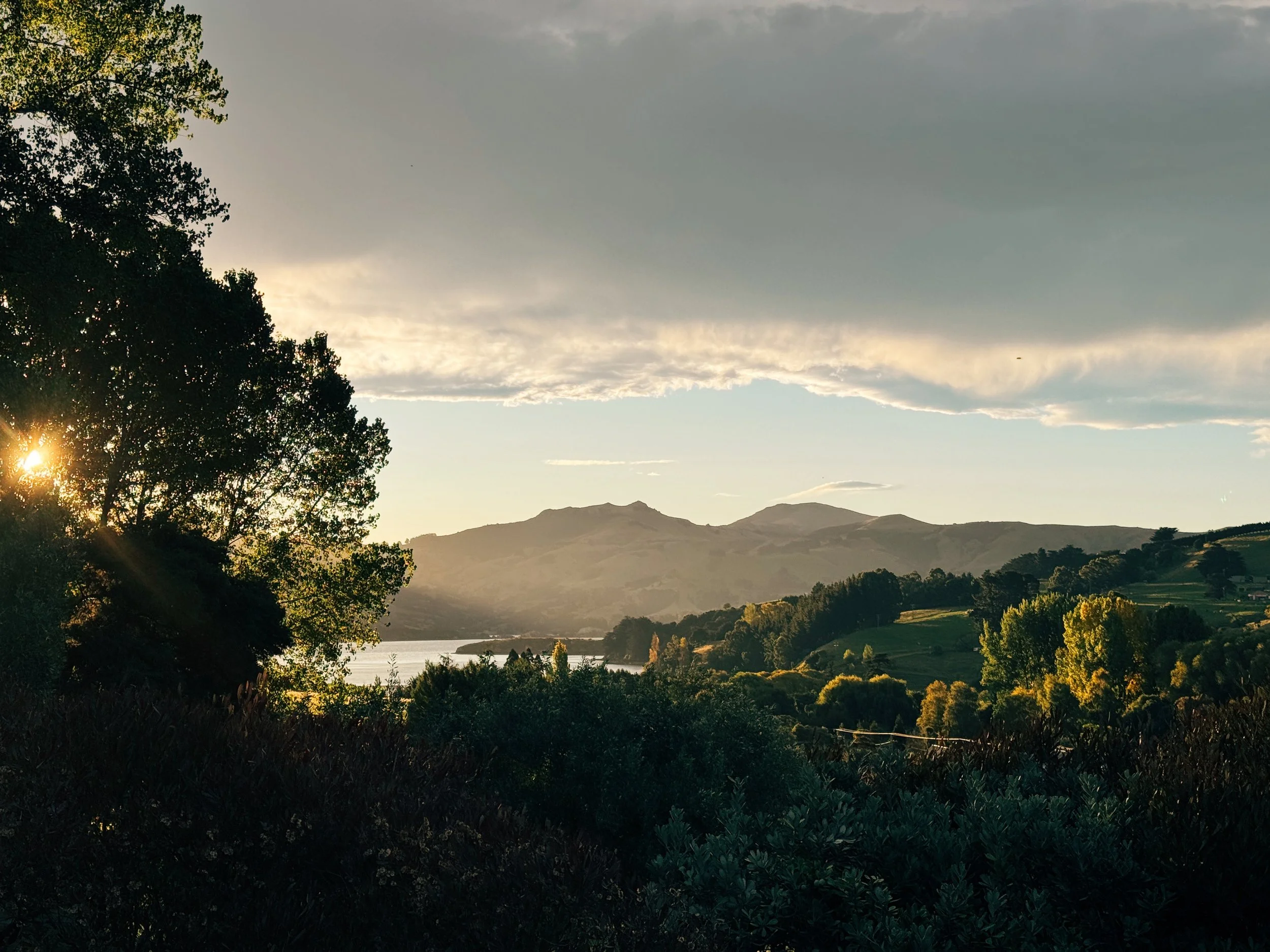 Sunset over rolling hills and mountains, with a lake and trees in the foreground.
