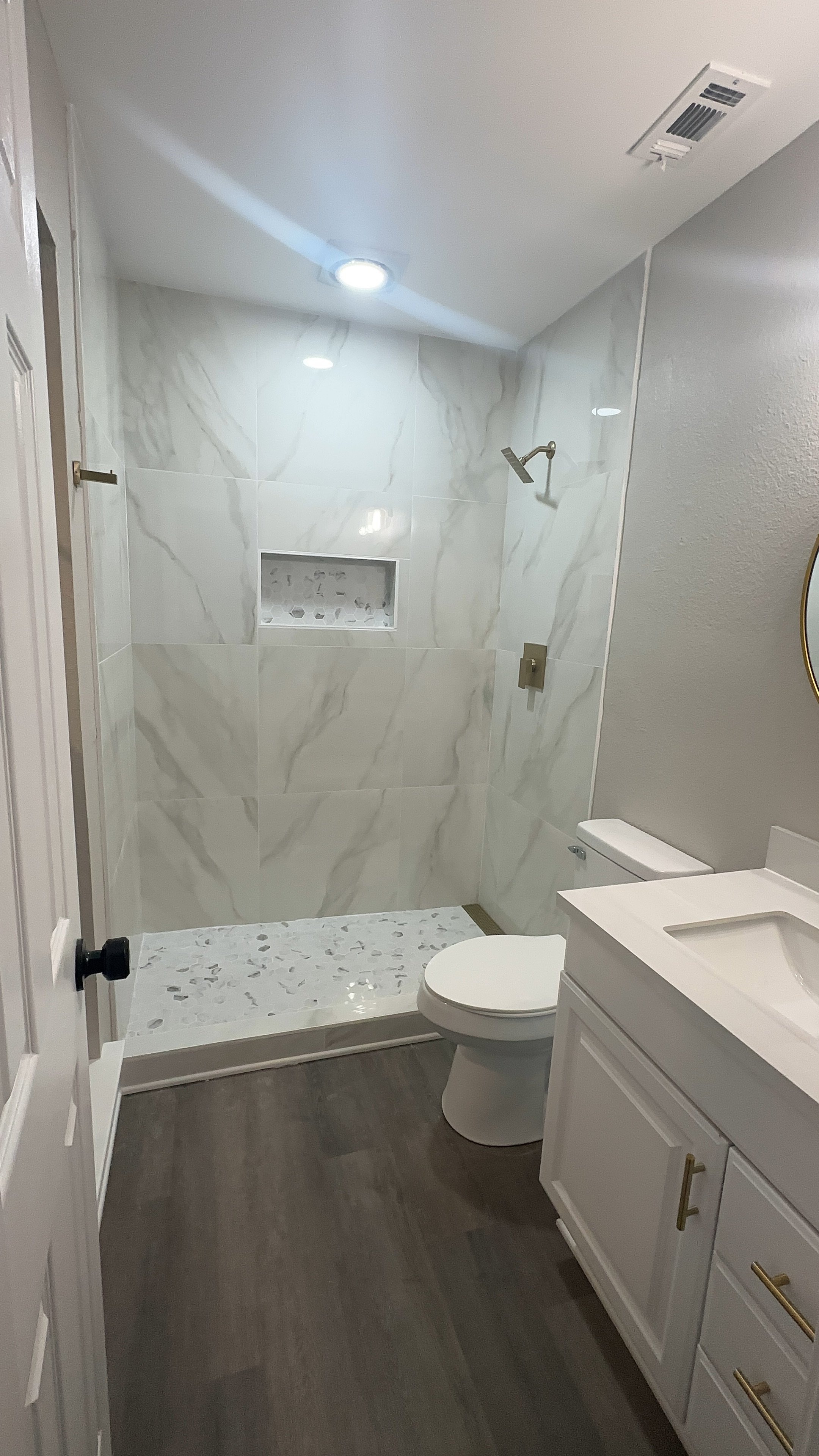 Modern bathroom with a walk-in shower, white vanity with gold hardware, toilet, dark wood floor, and gray walls. Post renovation