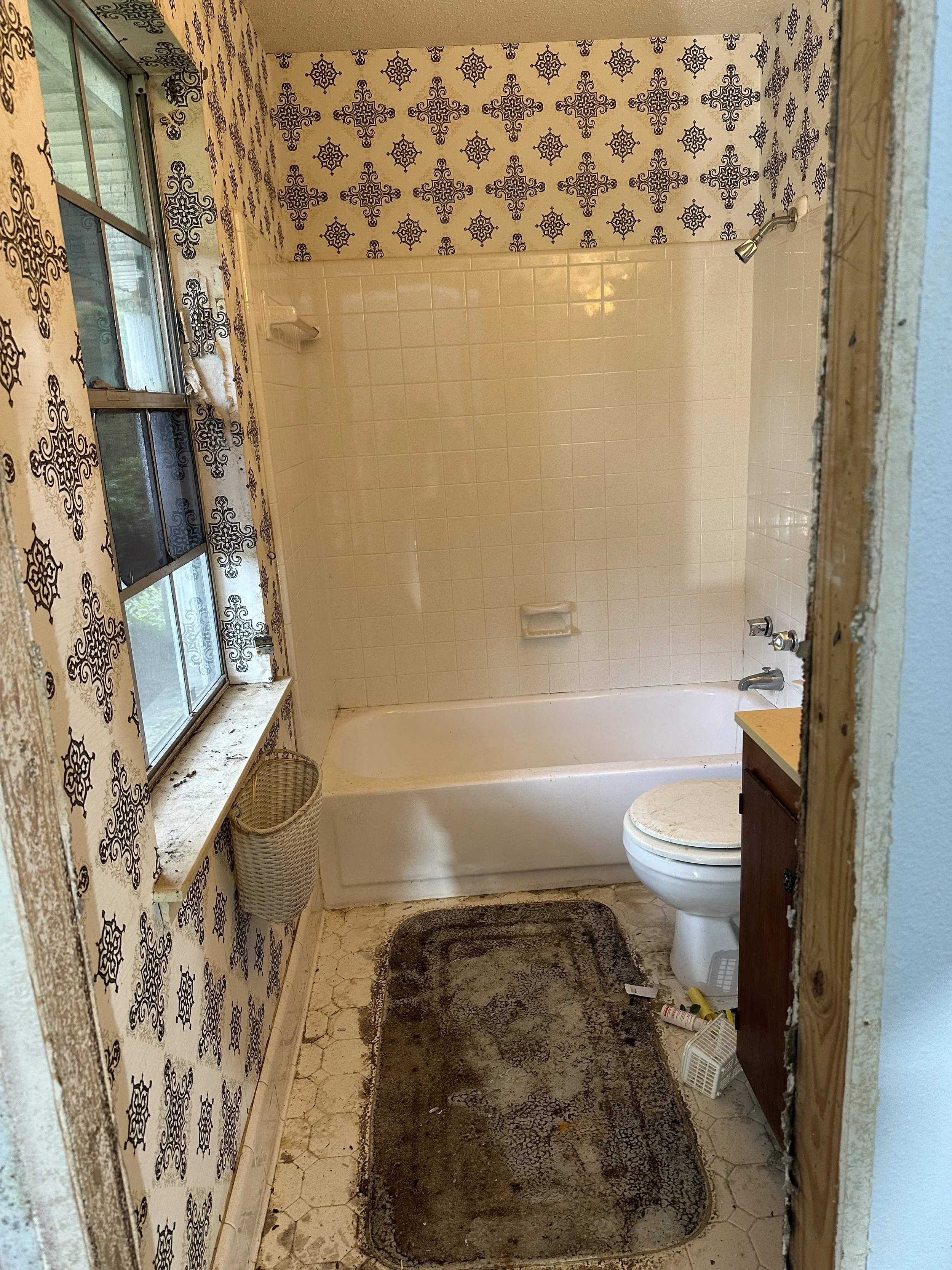 Original bathroom with a bathtub, toilet, window, and wallpaper with a black floral pattern. The floor has a stained, dark-colored rug, and the wall near the window has peeling wallpaper. Pre renovation