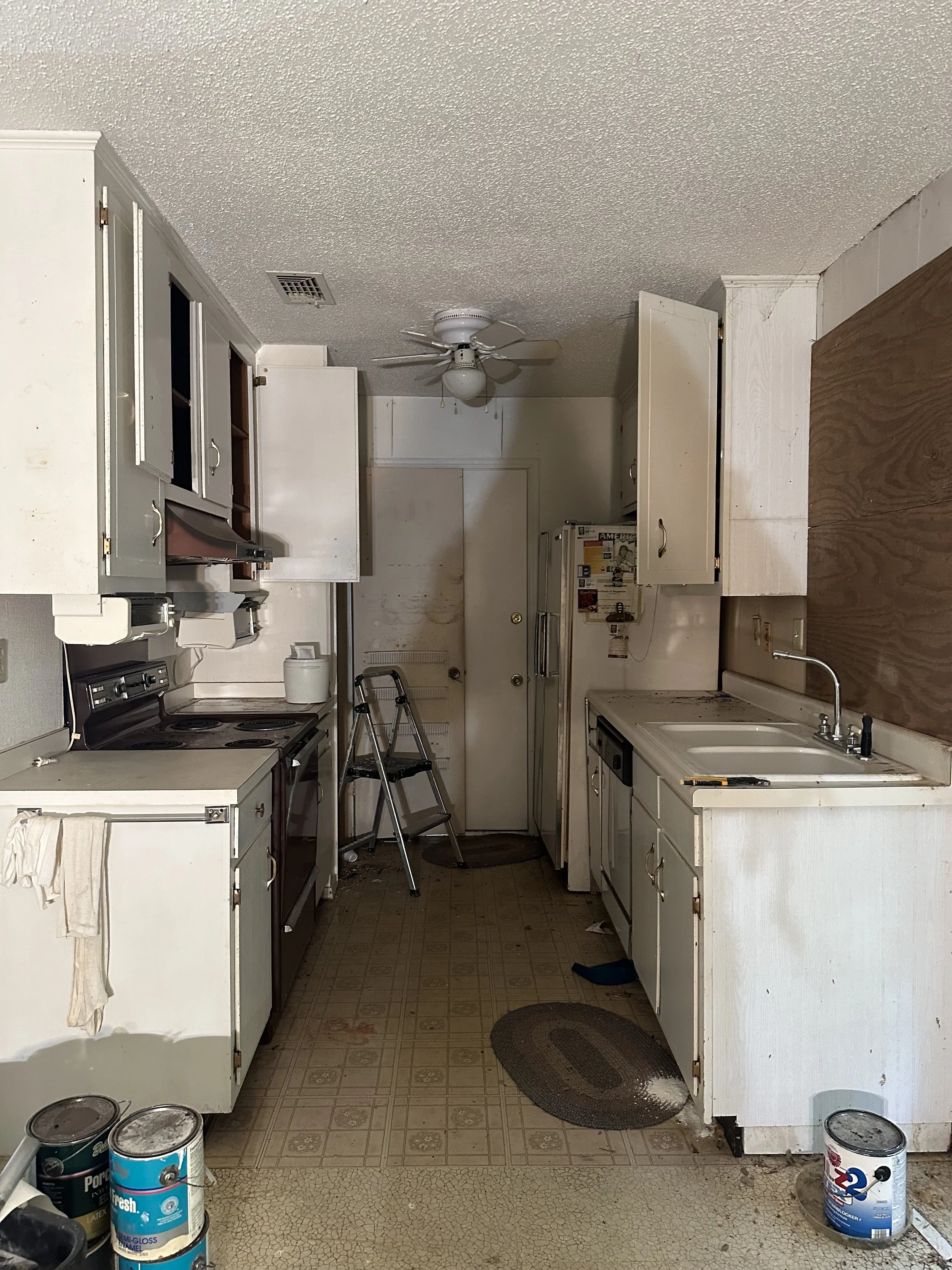 A cluttered kitchen with old white cabinets and a ceiling fan. The floor is worn with several paint cans on it. A step stool is in front of the door, and there are dirty spots on the walls and floor. Pre renovation