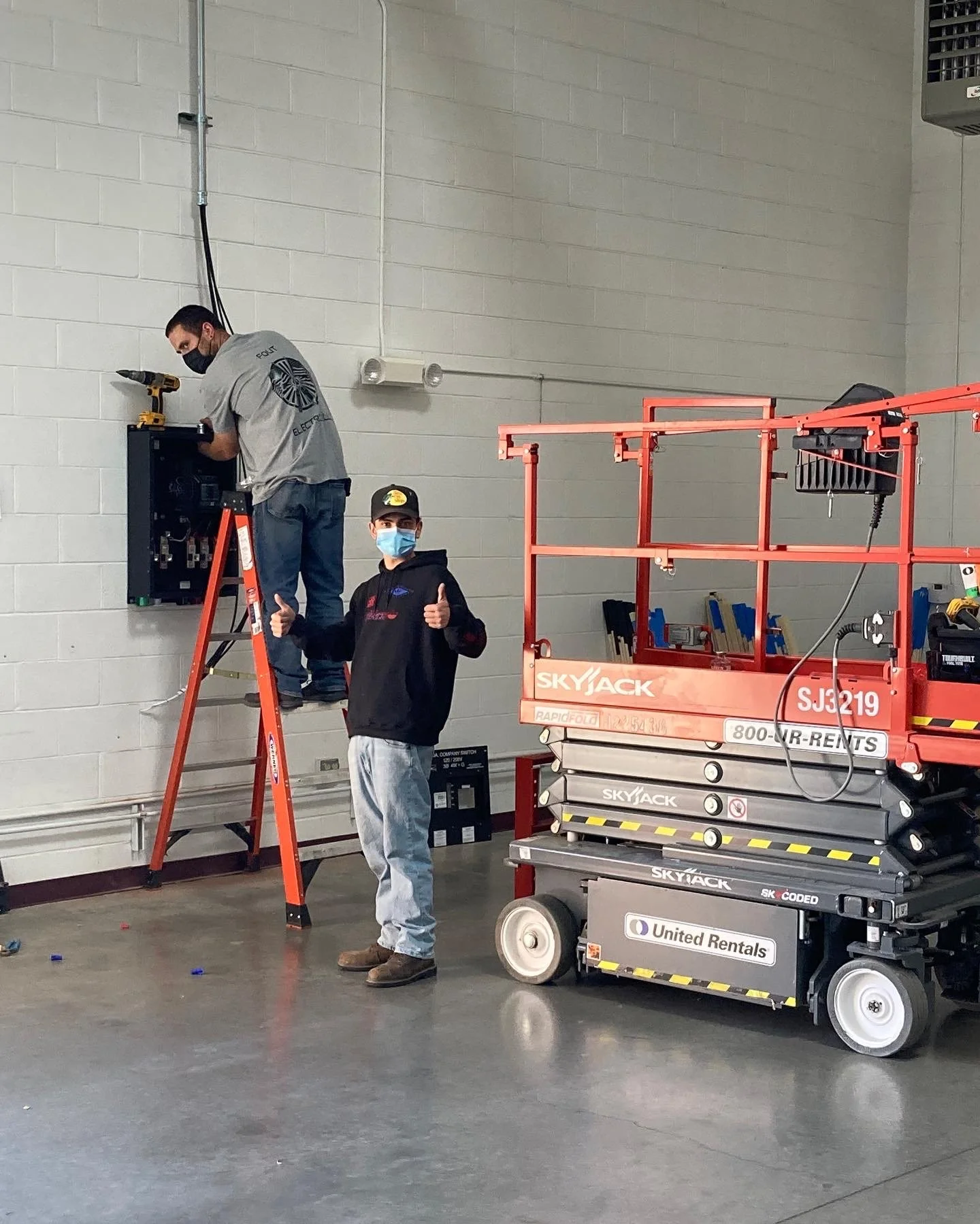 Two workers performing maintenance on electrical equipment. One worker is standing on the ground giving a thumbs-up, while the other is on a ladder working on an electrical panel. Both workers are wearing face masks. There is a red lift platform nearby and a scissor lift labeled with the logo 'Skyjack'.