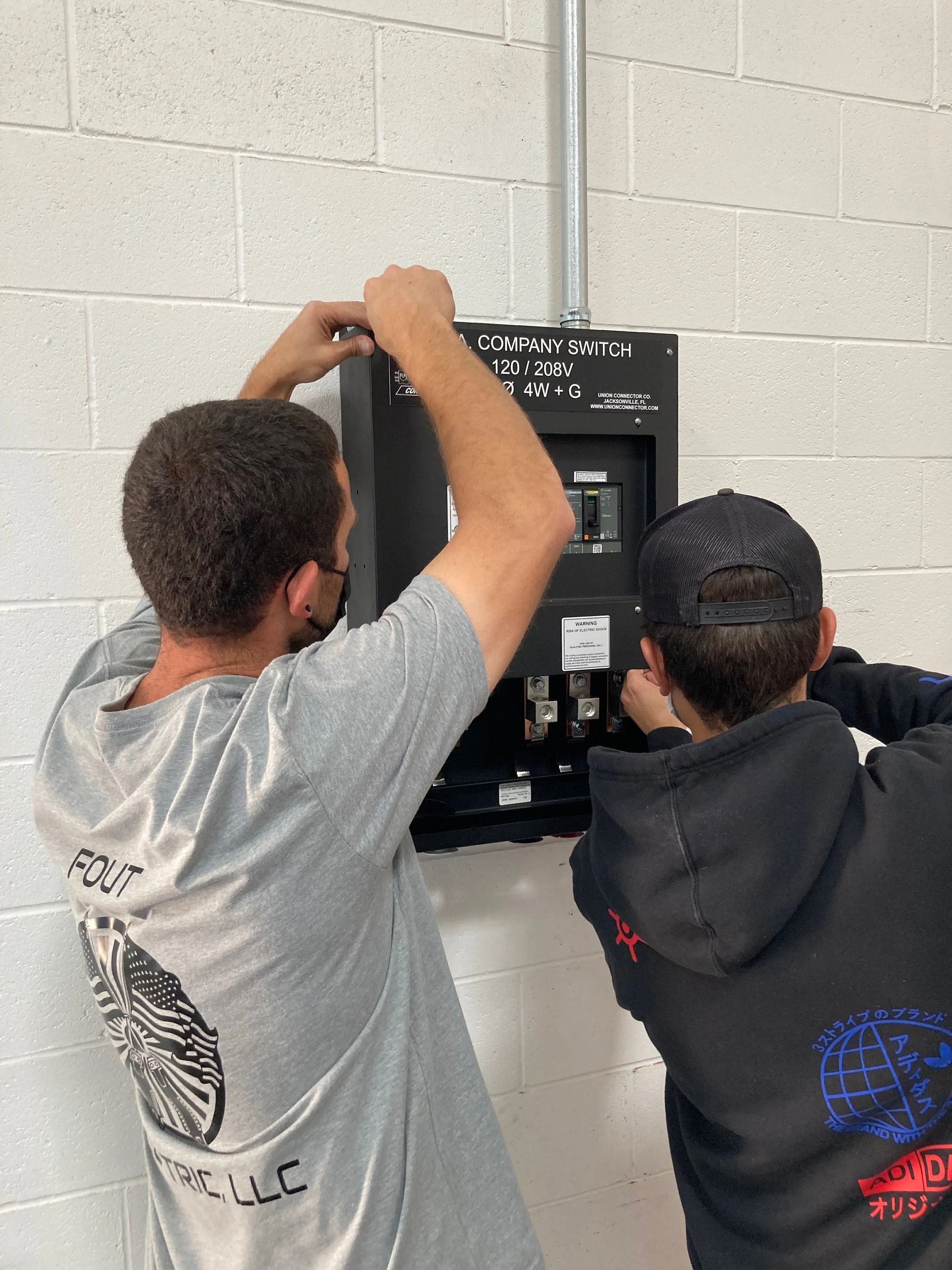 Two men working on an electrical panel labeled 'company switch' on a white brick wall.