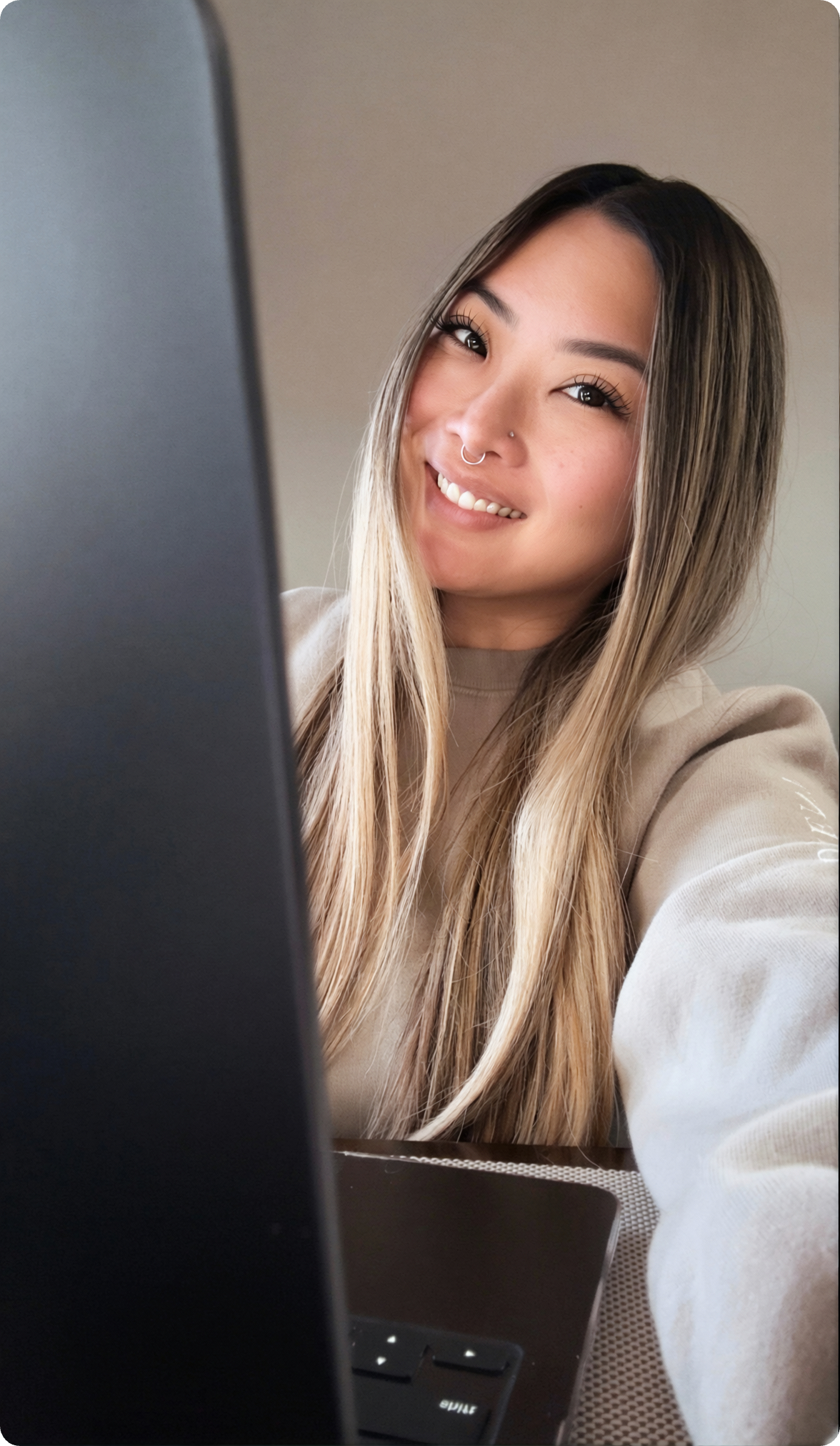 Young woman with long hair, wearing a beige sweatshirt, smiling while sitting at a desk with a laptop.