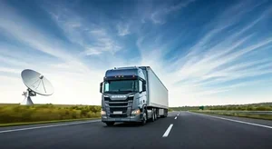 A large gray semi-truck driving on an open highway with a satellite dish in the background, under a blue sky with scattered clouds.